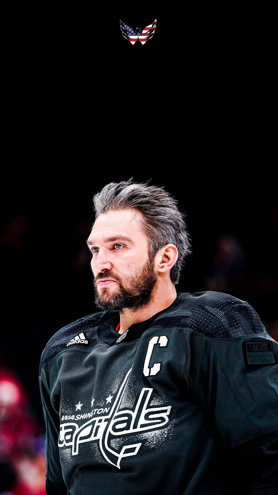 Washington Capitals 桌布 壁紙 — 一位專注的冰球隊隊長，身穿黑色華盛頓首都隊球衣，帶有隊長 'C'，在黑暗的競技場背景下凝視著。