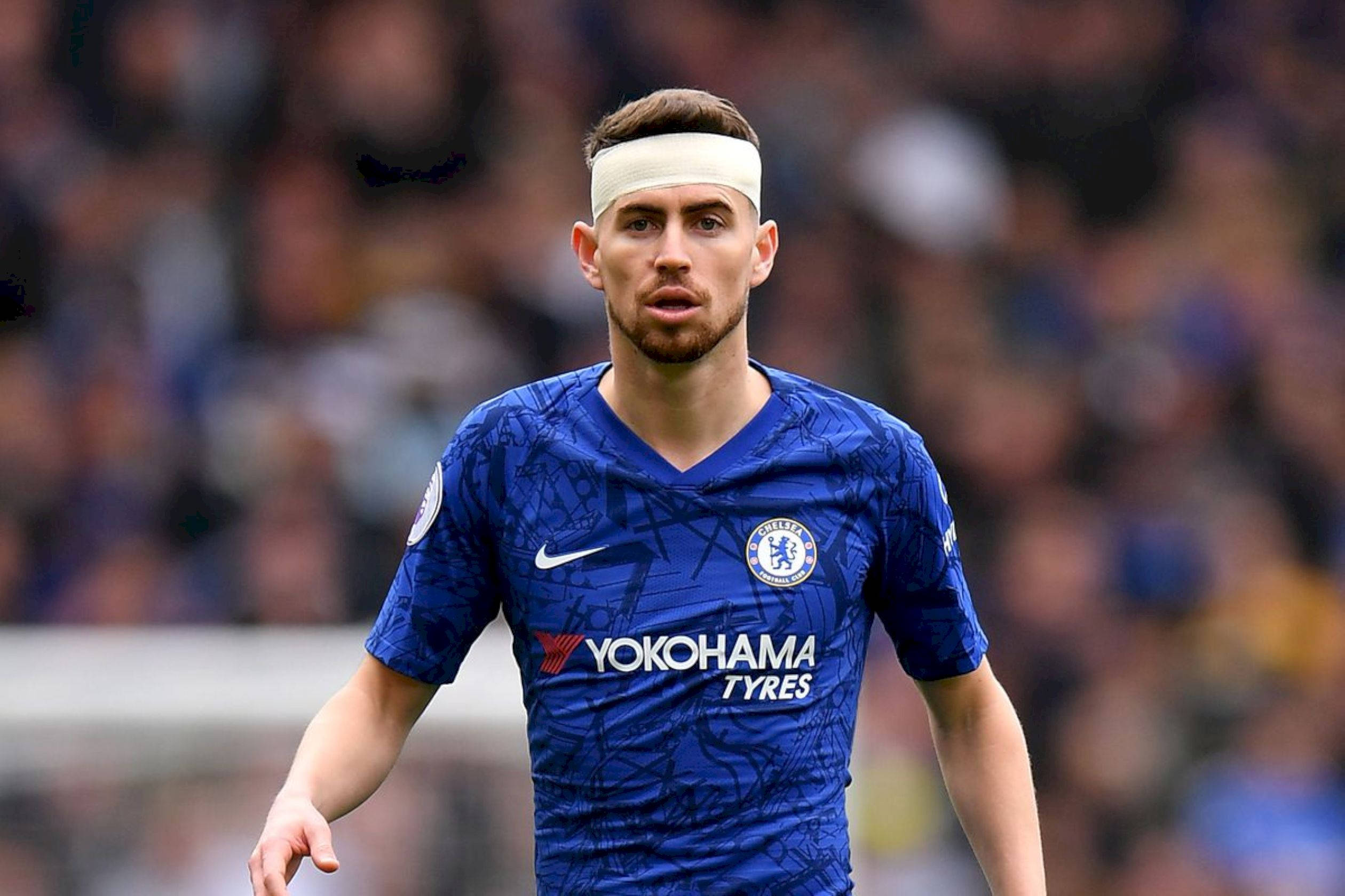 足球 桌布 壁紙 — 一位專注的切爾西足球員身穿藍色球衣、戴著白色頭帶，在陽光普照的球場上大步走過，背景是模糊的人群。