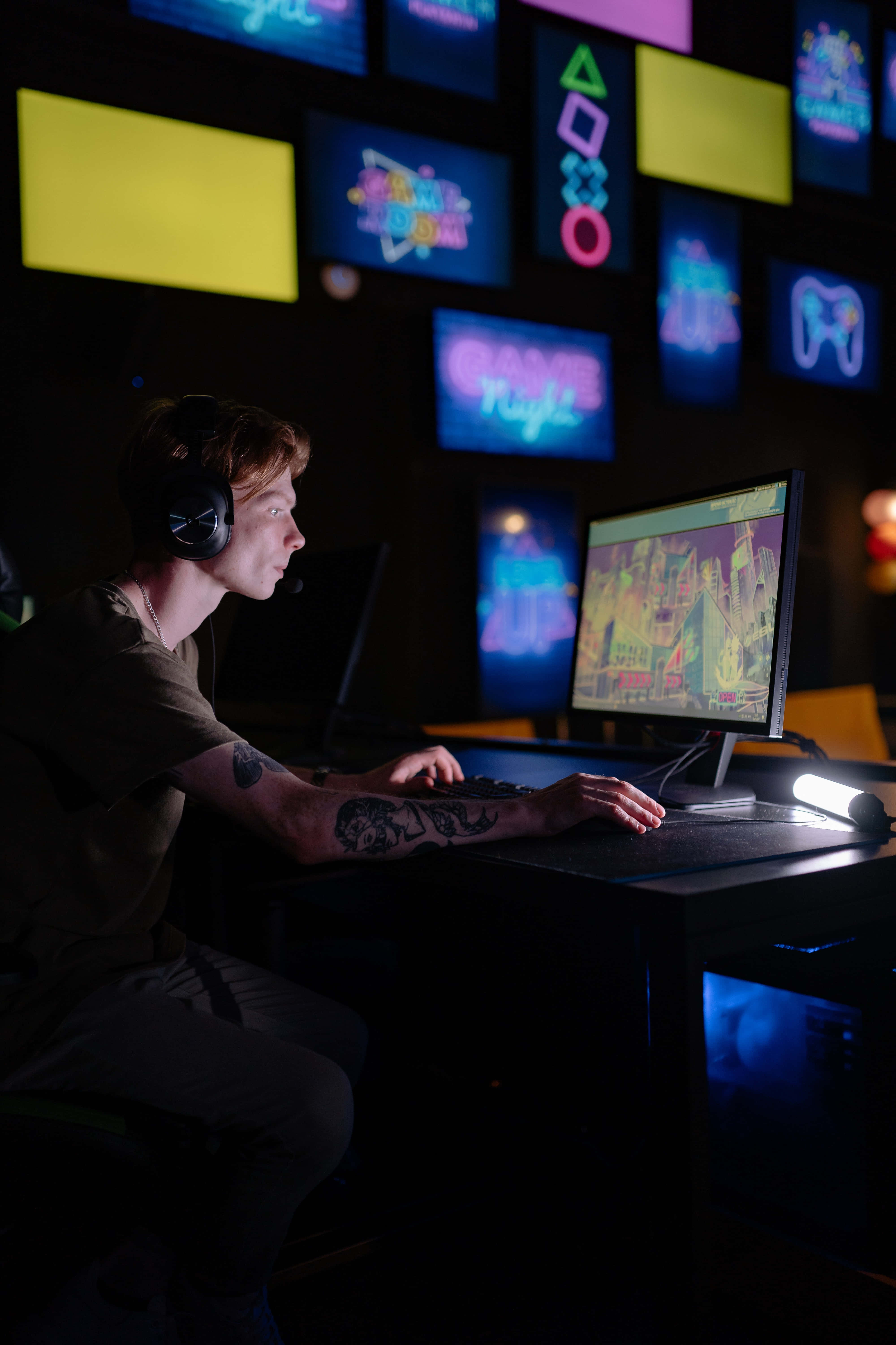 Gaming Room 桌布 壁紙 — 一位專注的刺青玩家在霓虹燈光下的街機主題牆飾前玩色彩繽紛的復古風格遊戲。