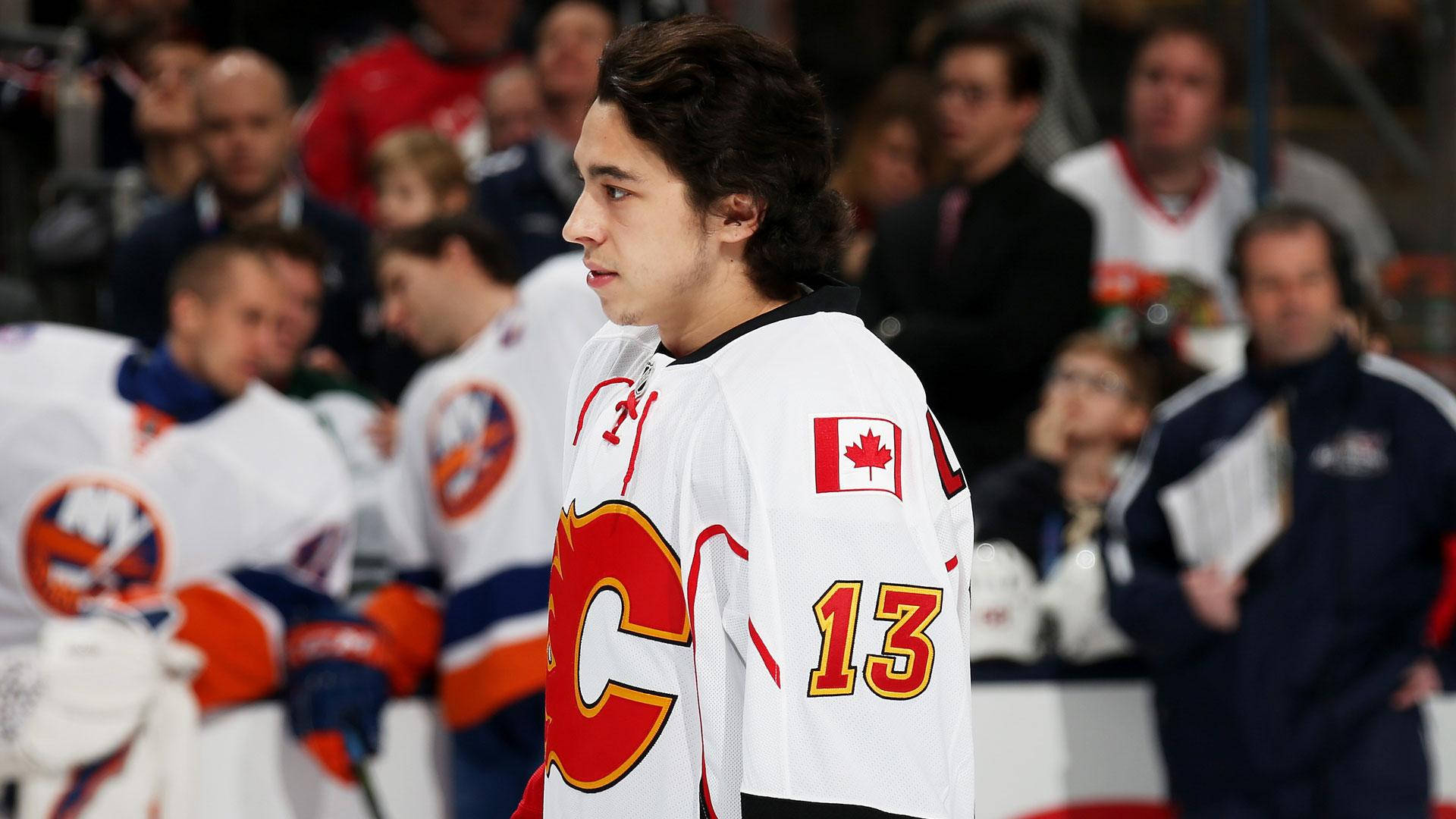 Johnny Gaudreau 桌布 壁紙 — 一位專注的加拿大曲棍球運動員，身穿白色球衣，上有紅色楓葉和13號，在比賽中站在冰上，背景是模糊的觀眾。