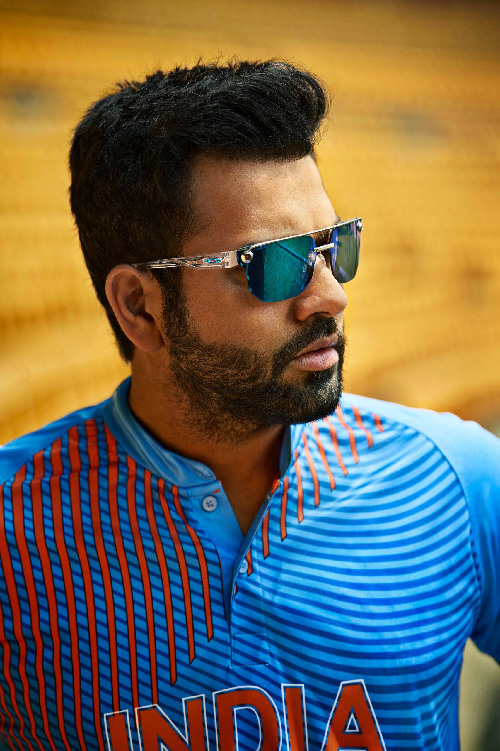 Sunglasses 桌布 壁紙 — 一位專注的印度板球運動員，身穿藍紅條紋球衣，戴著反光太陽眼鏡，背景是模糊的金色。