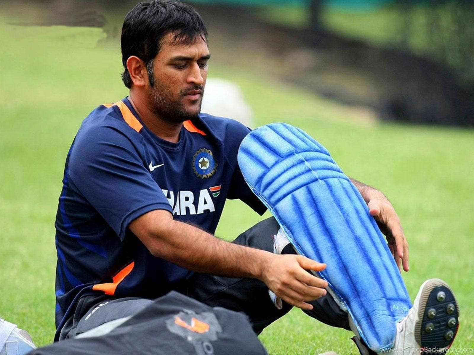 MS Dhoni 桌布 壁紙 — 一位專注的印度板球運動員坐在草地上，在訓練期間調整他的藍色護腿。
