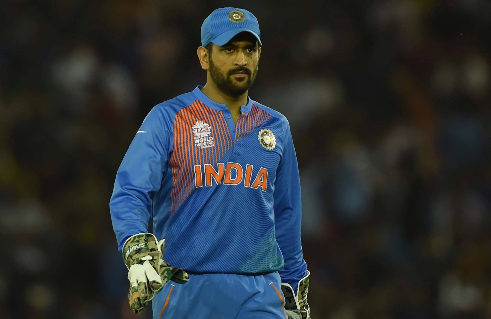 MS Dhoni 桌布 壁紙 — 一位專注的印度板球運動員穿著藍色制服，在夜燈下的體育場場地上莊嚴地行走。