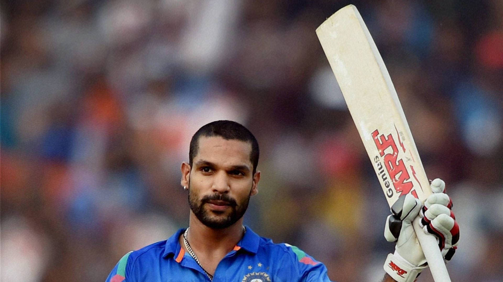 shikhar dhawan 桌布 壁紙 — 一位專注的印度板球運動員身穿藍色球衣，在模糊的體育場人群中高舉白色球棒。