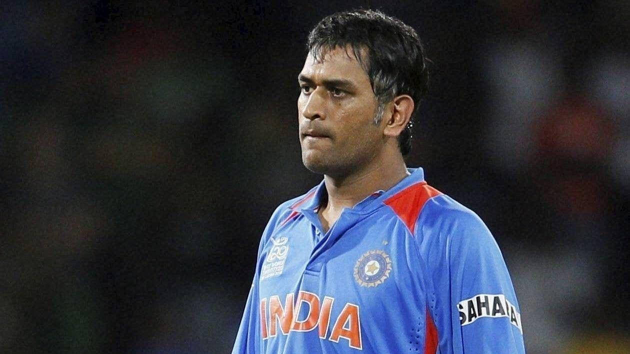 Ms Dhoni 桌布 壁紙 — 一位專注的印度板球運動員身穿藍色球衣，站在體育場燈光下，背景是模糊的人群。