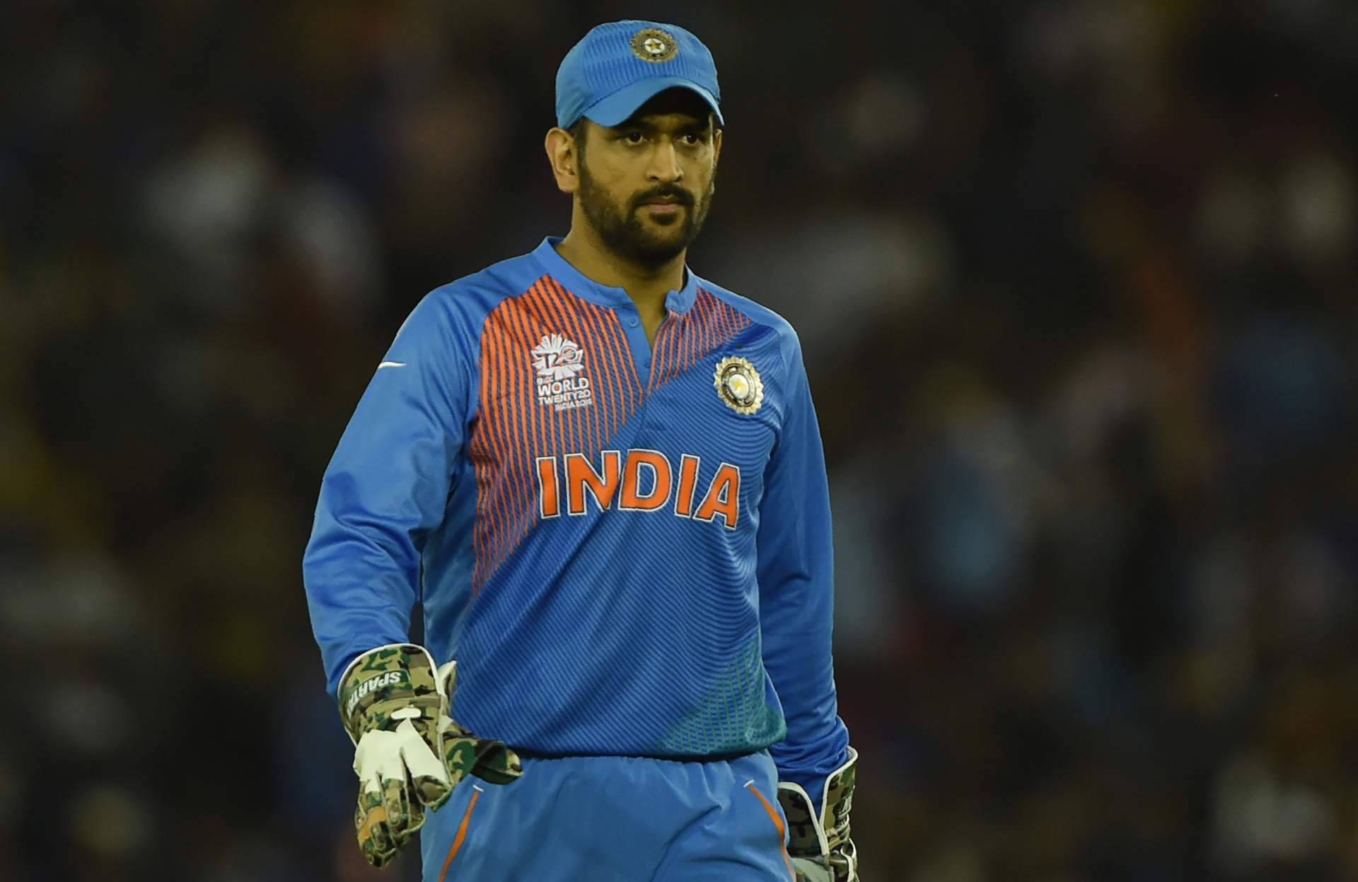 Ms Dhoni 桌布 壁紙 — 一位專注的印度板球運動員身穿藍色制服，在體育場燈光下莊嚴地走在體育場場地上。