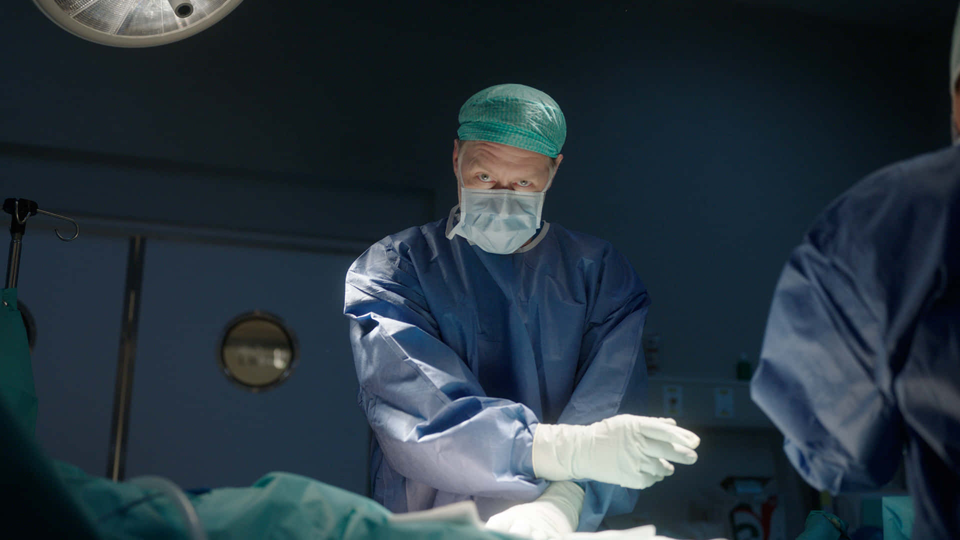 Hospital 桌布 壁紙 — 一位專注的外科醫生穿著藍色手術服和綠色帽子，在無菌手術室明亮的頂燈下進行手術。