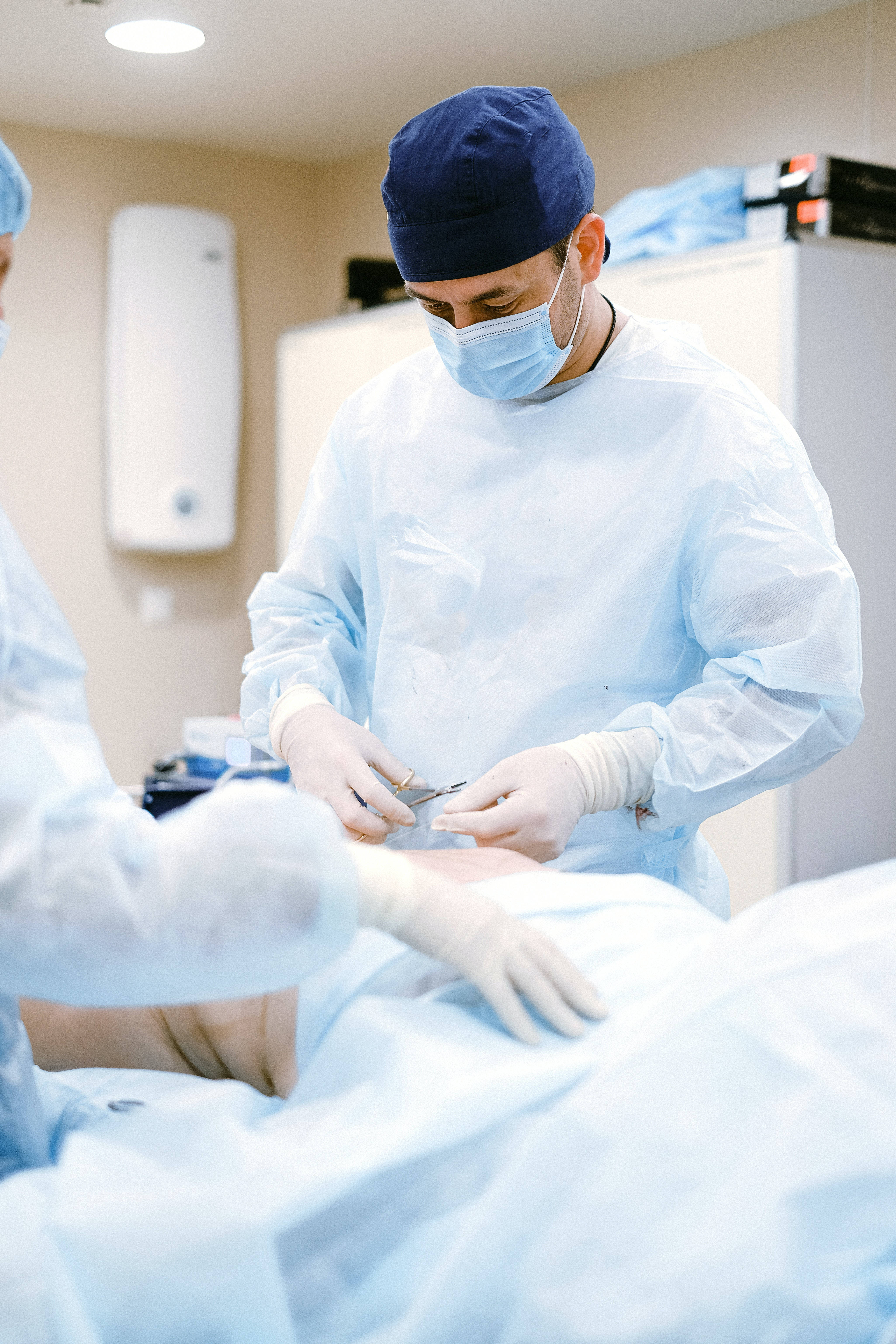 Surgeon 桌布 壁紙 — 一位專注的外科醫生穿著藍色手術衣和口罩，在明亮的手術燈光下進行精細的手術。