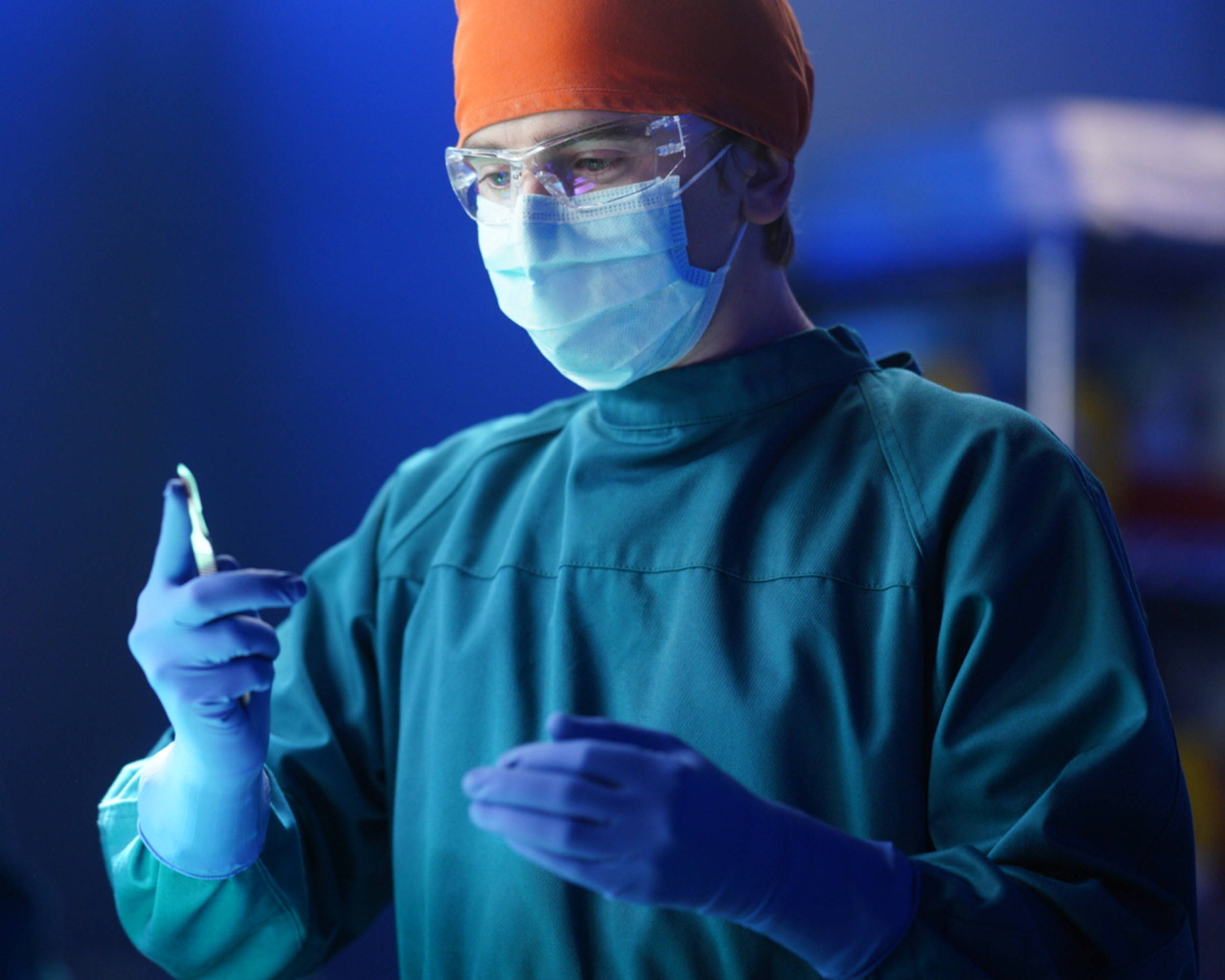 The Good Doctor 桌布 壁紙 — 一位專注的外科醫生穿著青色手術服和橙色帽子，在冷藍色的手術室燈光下拿著手術器械。