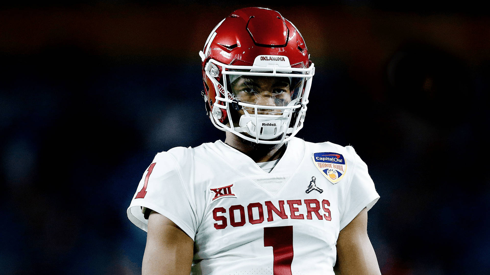 運動 桌布 壁紙 — 一位專注的奧克拉荷馬大學 Sooners 美式足球員，身穿白色和紅色裝備，在體育館燈光下凝視。