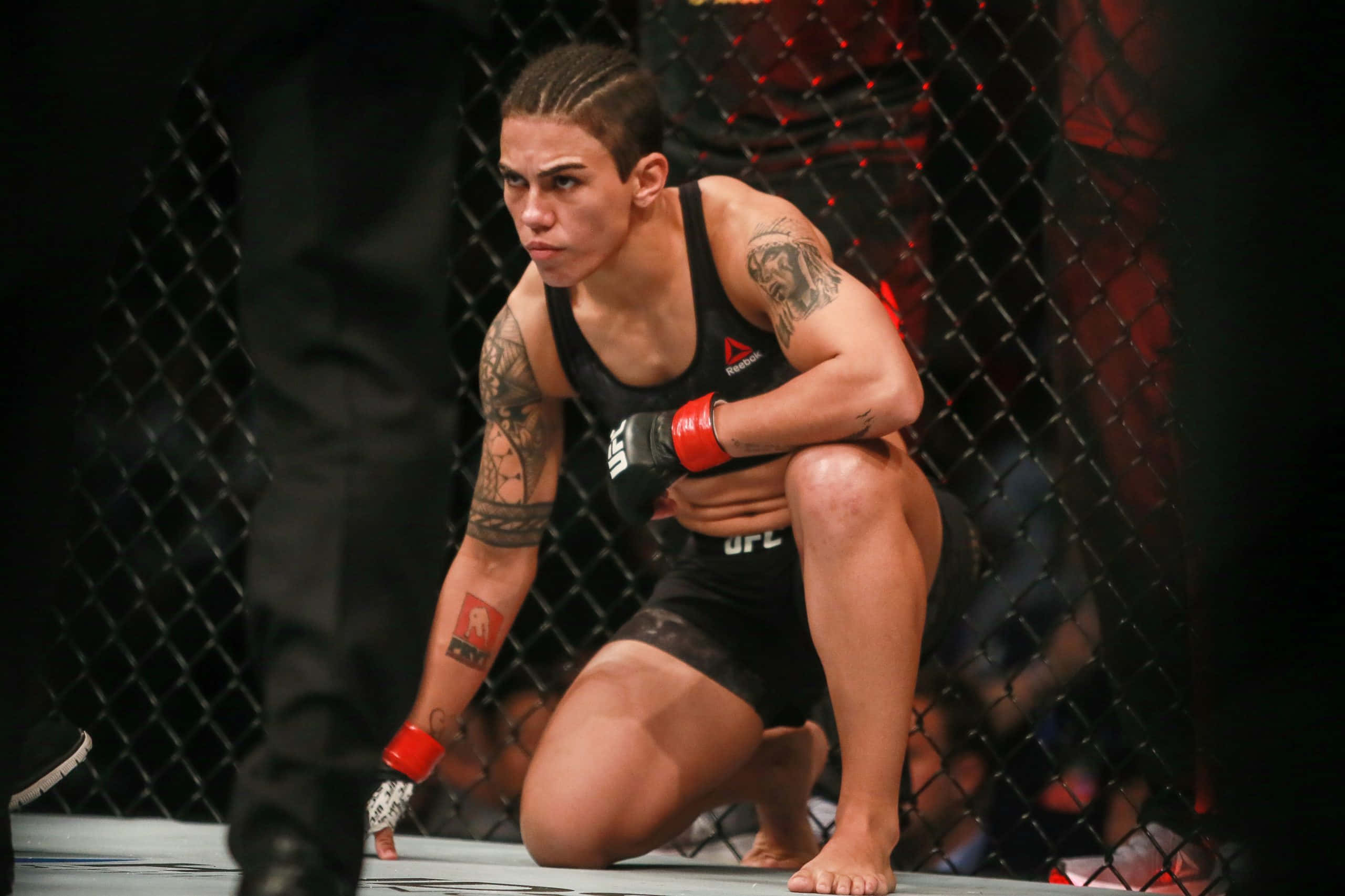 MMA 桌布 壁紙 — 一位專注的女性MMA拳手在籠內跪下，身穿黑色裝備和紅色手套，手臂上可見刺青。