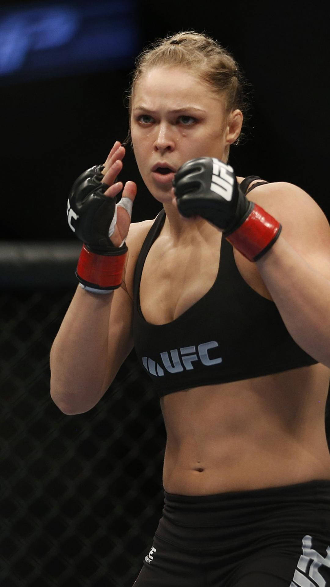 Rousey 桌布 壁紙 — 一位專注的女性MMA拳手穿著黑色UFC裝備在昏暗的八角籠中出拳。
