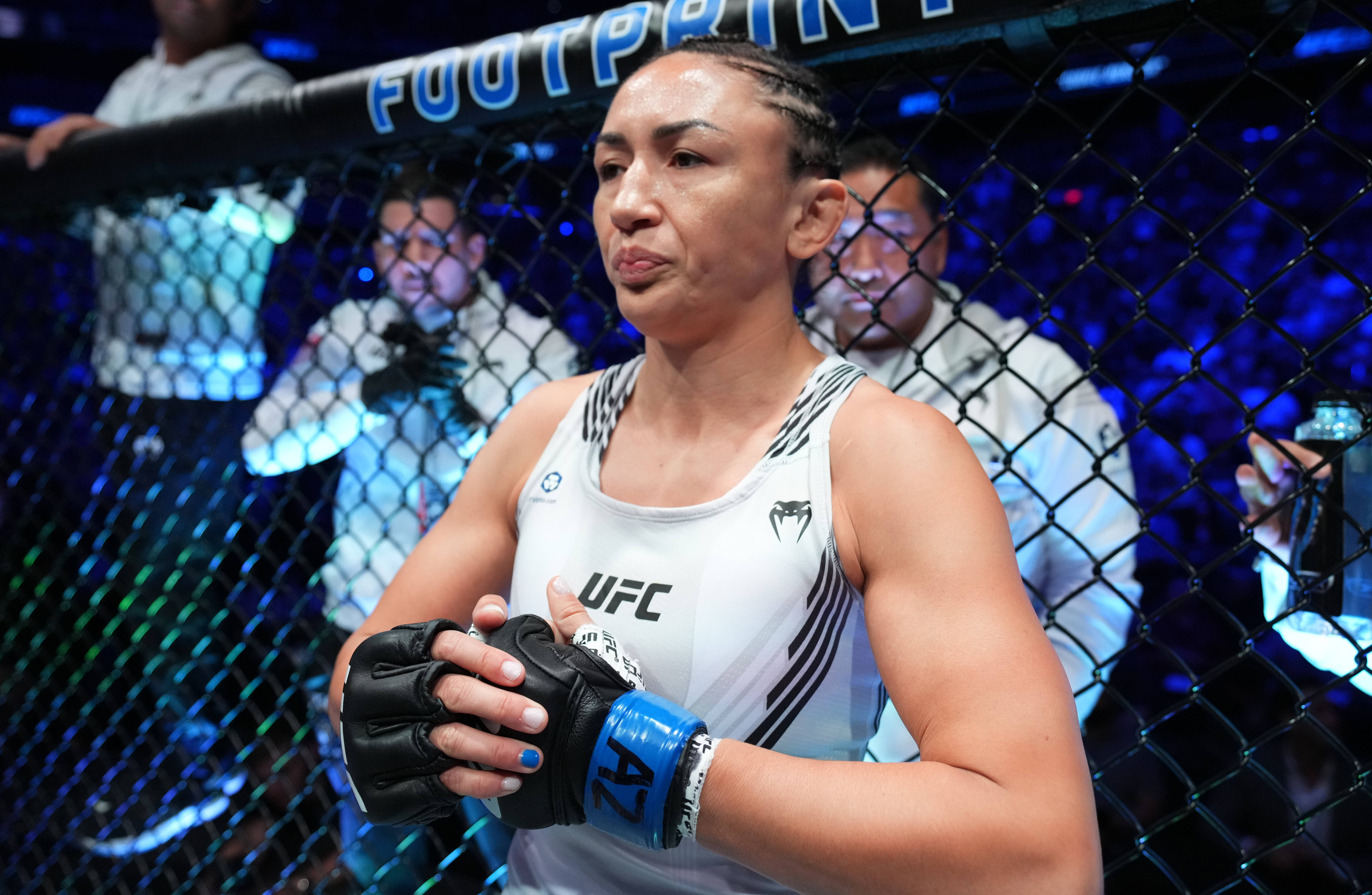 MMA 桌布 壁紙 — 一位專注的女性MMA拳手身穿白色UFC背心，站在藍色燈光的八角籠中，雙手交握。