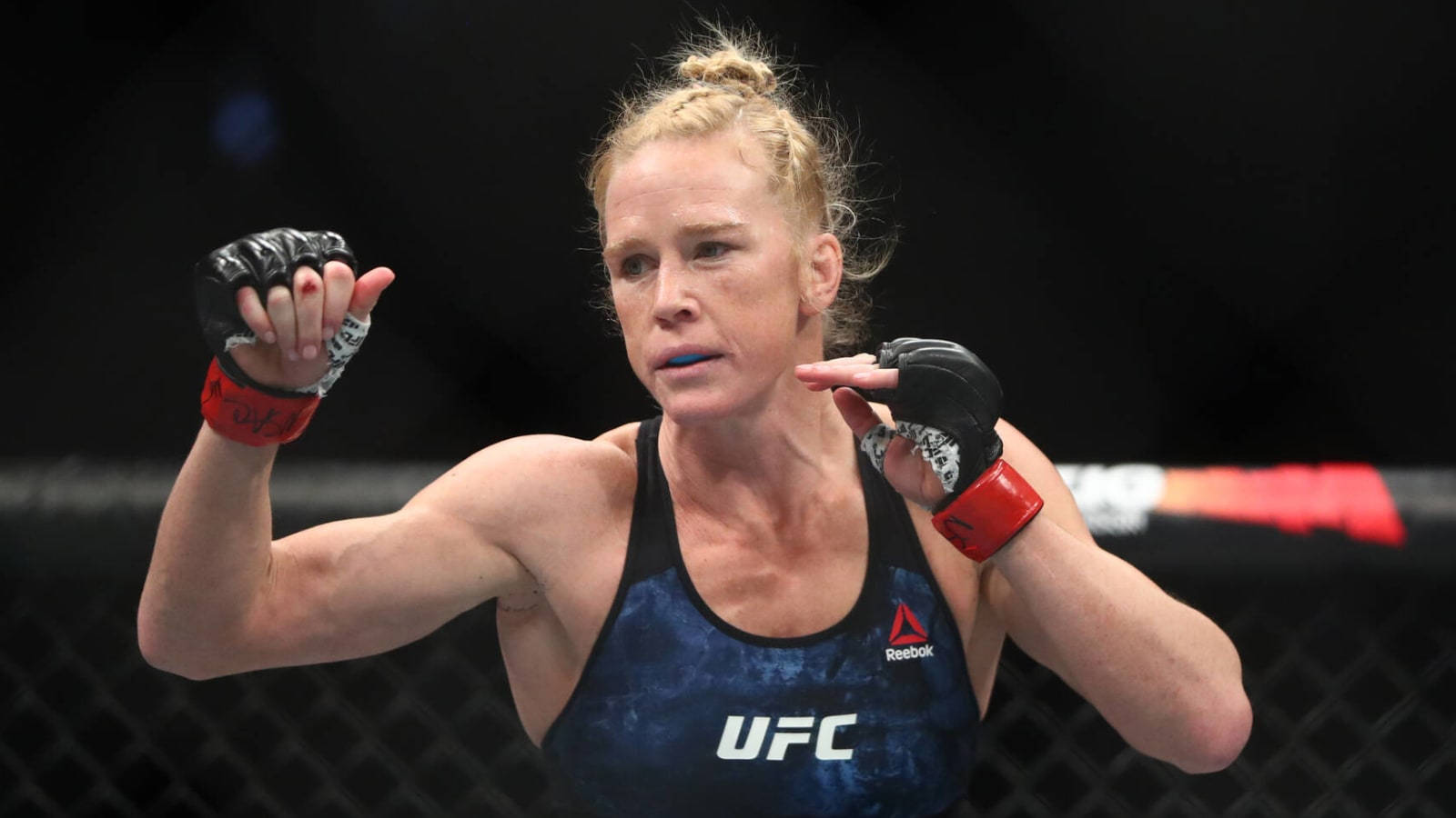 Fight 桌布 壁紙 — 一位專注的女性MMA拳擊手，身穿藍色UFC背心和紅色手套，在黑暗的八角籠中準備就緒。