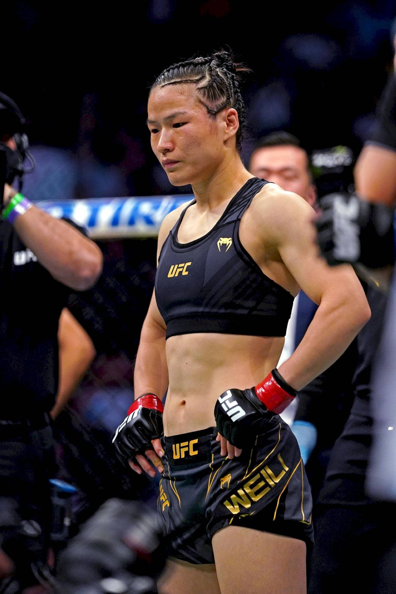 Athletic 桌布 壁紙 — 一位專注的女性UFC拳手，身穿黑色和金色裝備，在比賽後帶著堅定的表情站在八角籠中。