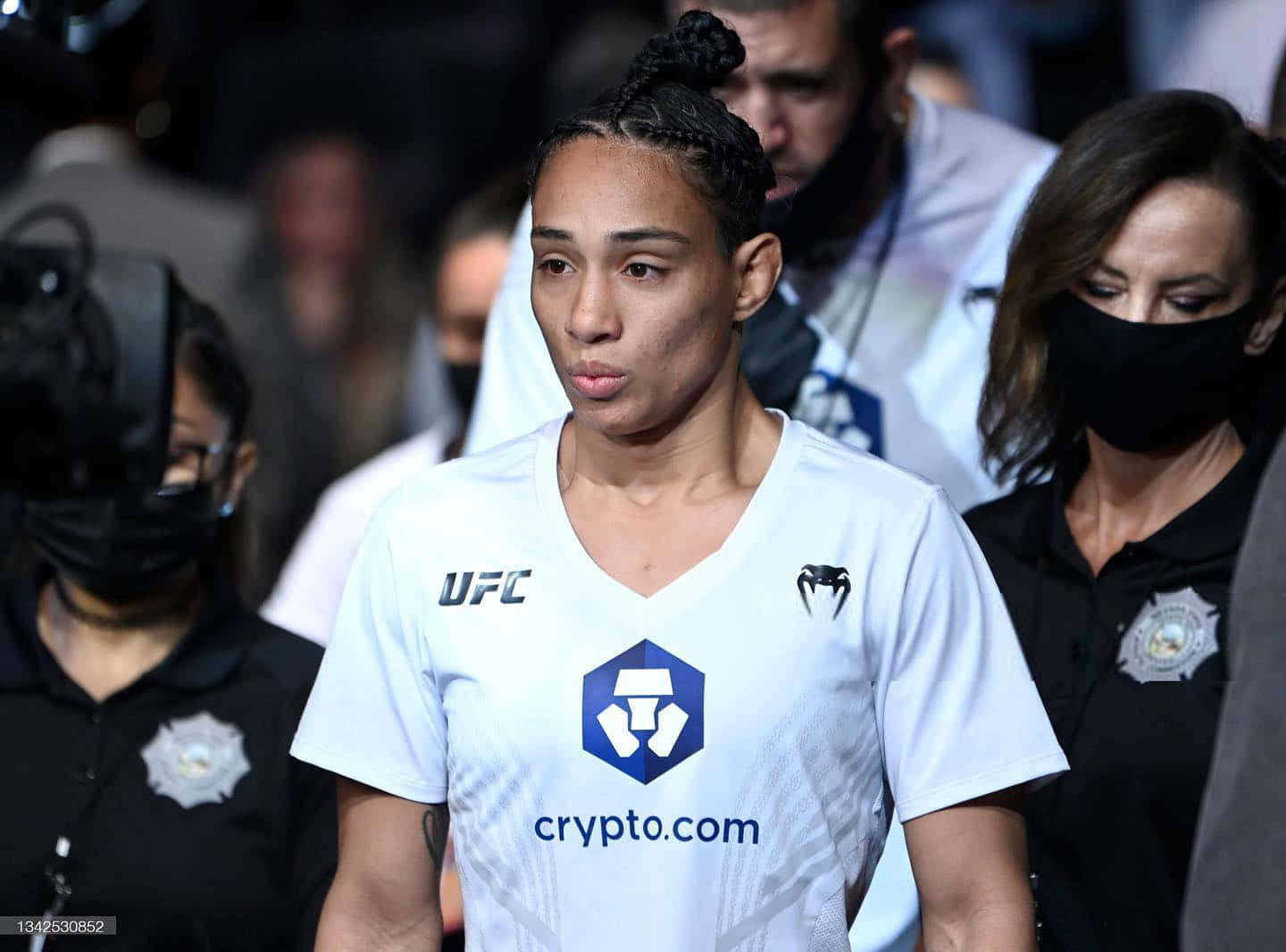 UFC 桌布 壁紙 — 一位專注的女性UFC拳手穿著白色crypto.com球衣，在保安人員陪同下穿過擁擠的場館。