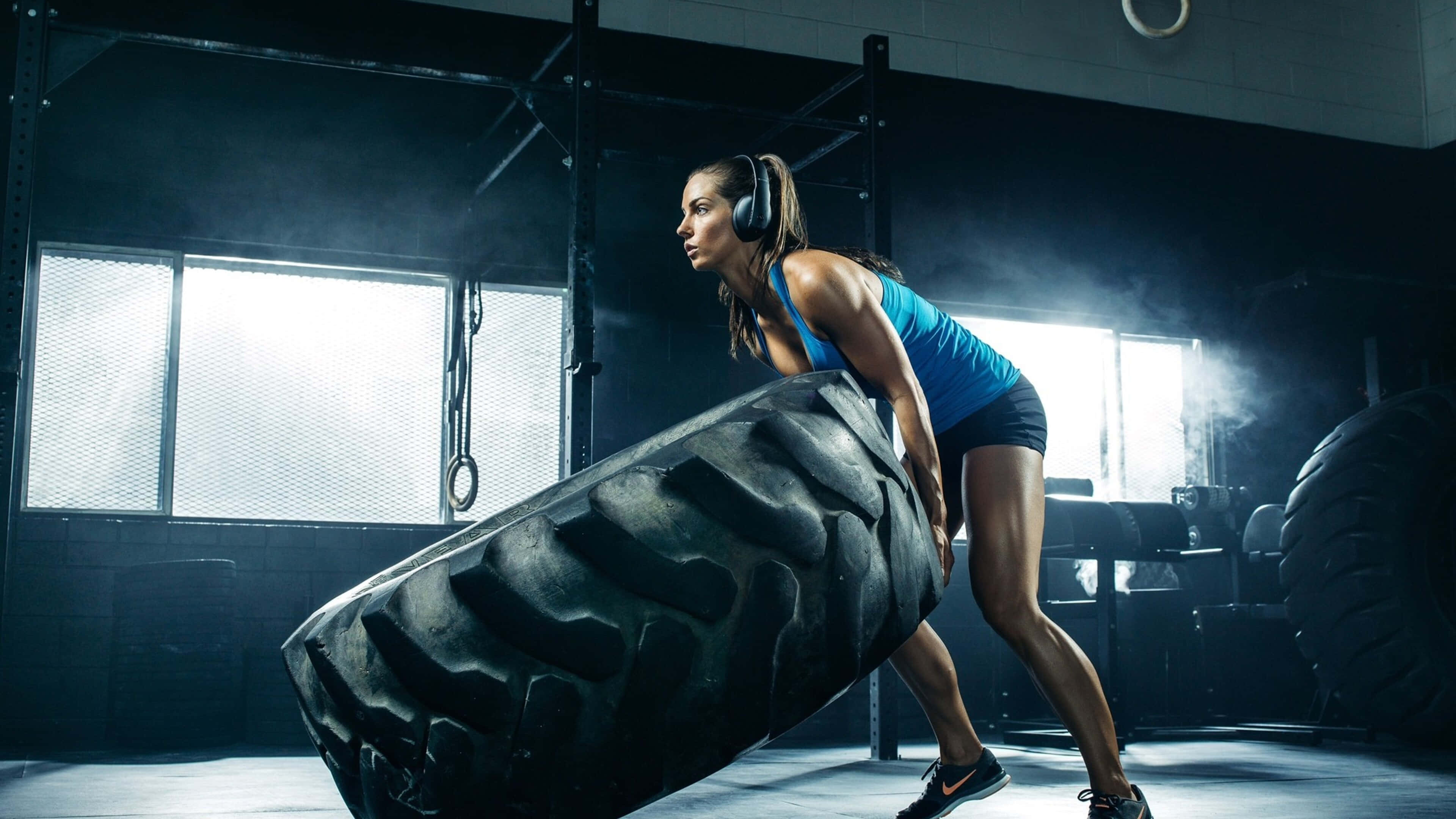 Fitness 桌布 壁紙 — 一位專注的女性在健身房中，在戲劇性的燈光下，以強烈的決心舉起一個巨大的黑色輪胎。