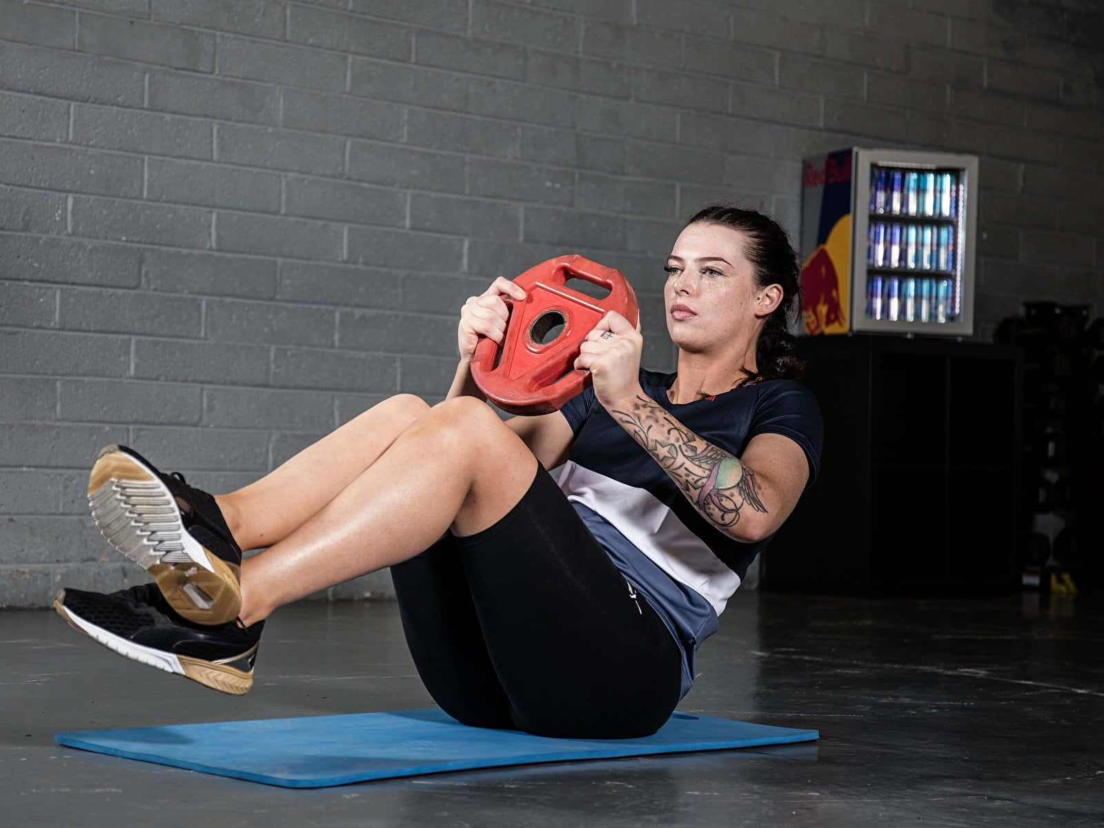 Workout 桌布 壁紙 — 一位專注的女性在健身房的灰色磚牆前，於藍色墊子上用紅色舉重片進行核心訓練。