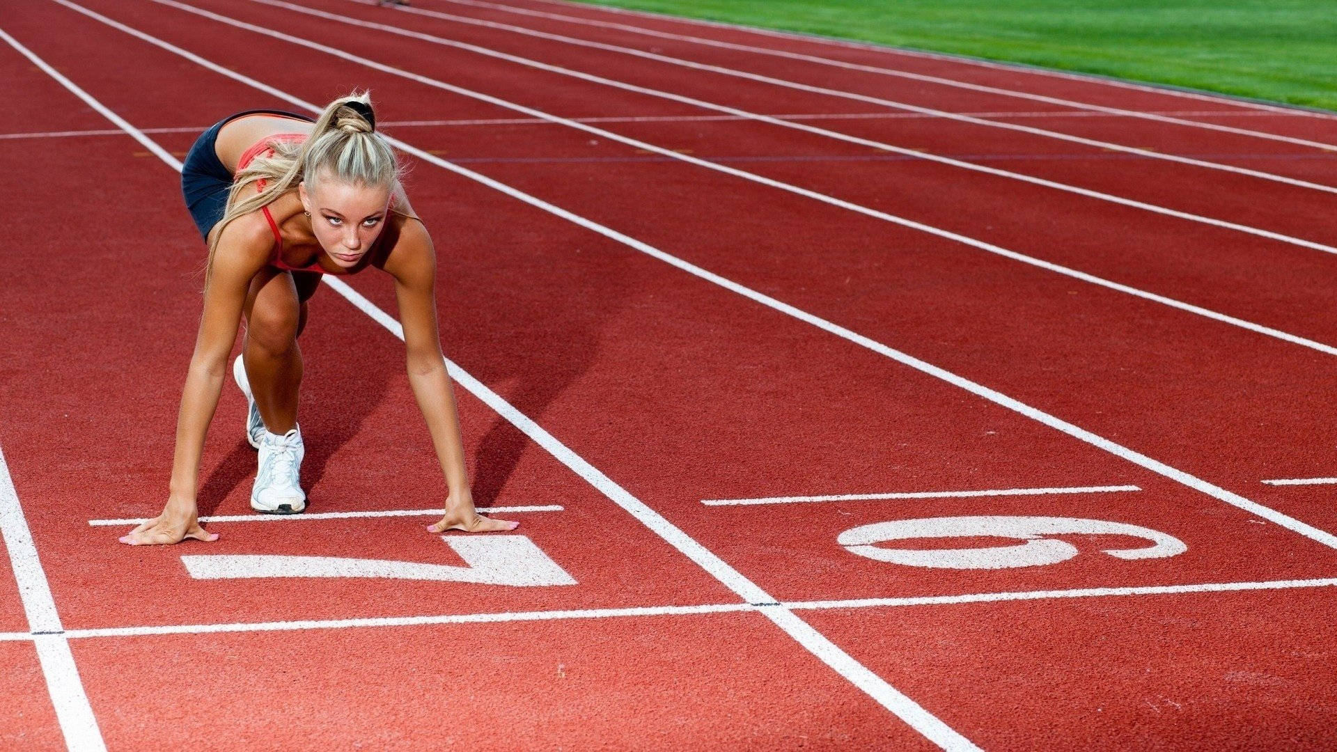 Athletic 桌布 壁紙 — 一位專注的女性短跑選手在紅色跑道上的起跑位置，跑道上有白色線條標記，在明亮的日光下。