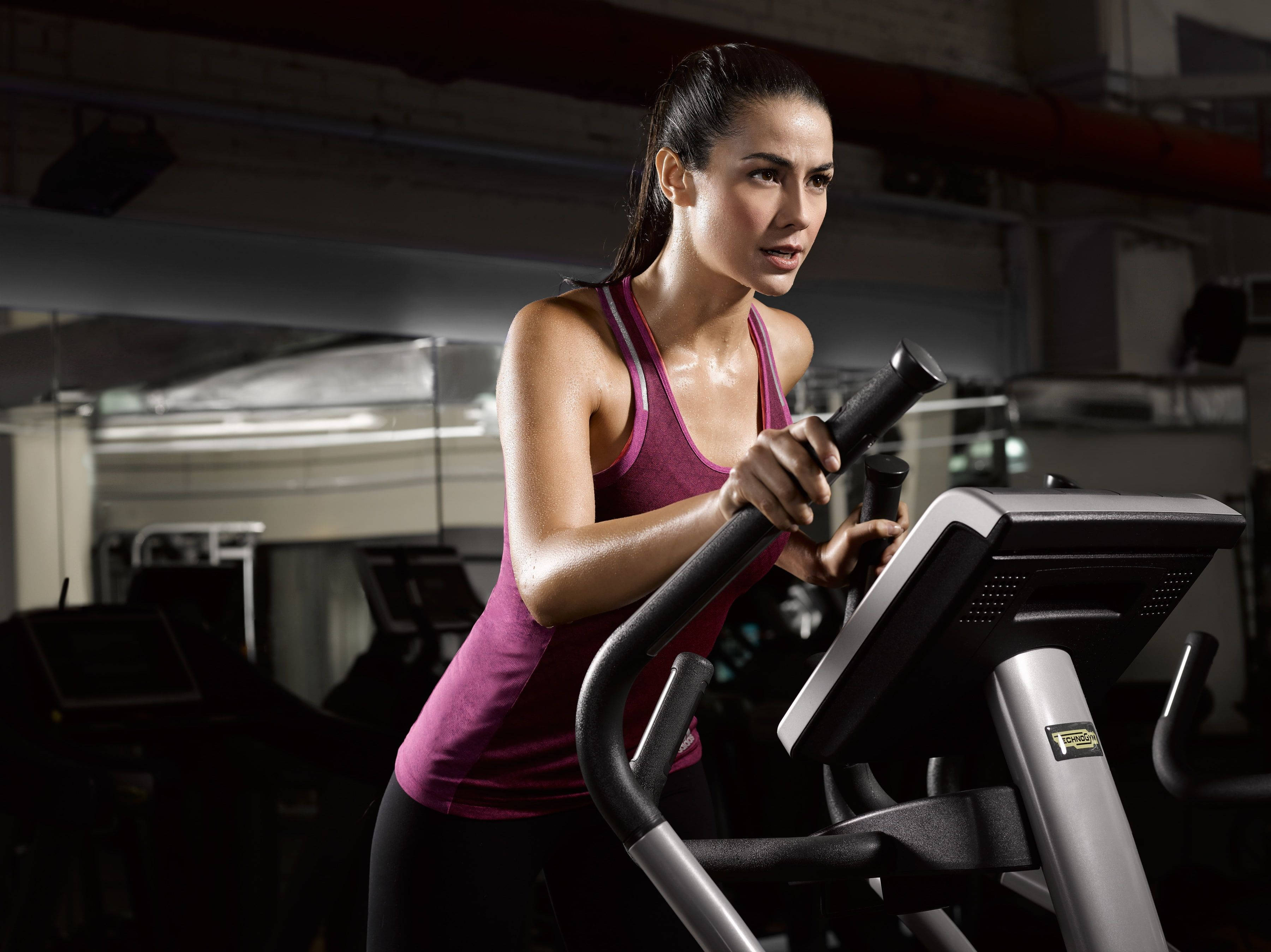Exercise 桌布 壁紙 — 一位專注的女性穿著紫色背心，在昏暗的健身房中於橢圓機上劇烈運動。