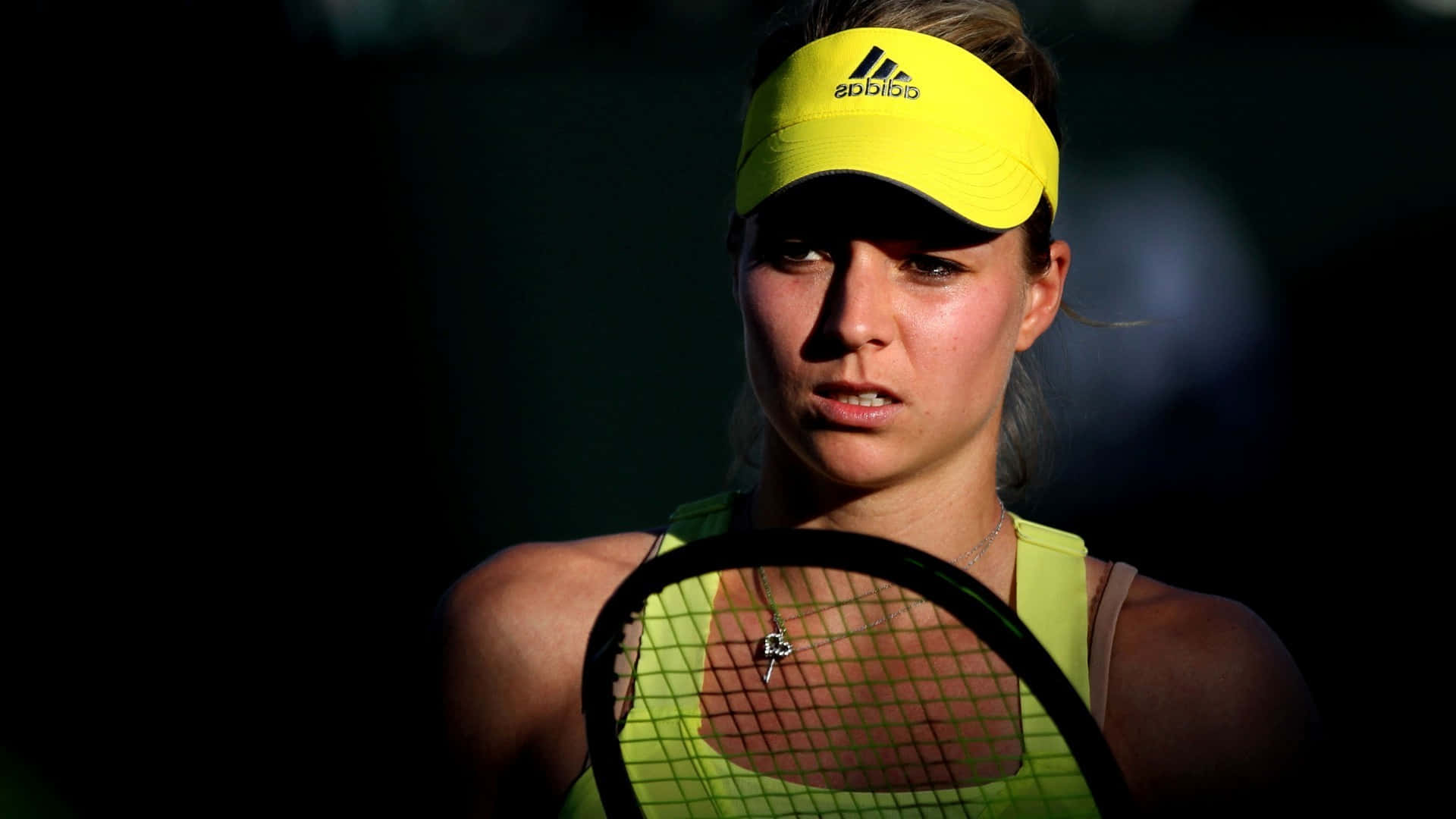HD Tennis 桌布 壁紙 — 一位專注的女性網球運動員，戴著亮黃色遮陽帽，身穿螢光綠色上衣，在深色背景下握著球拍。