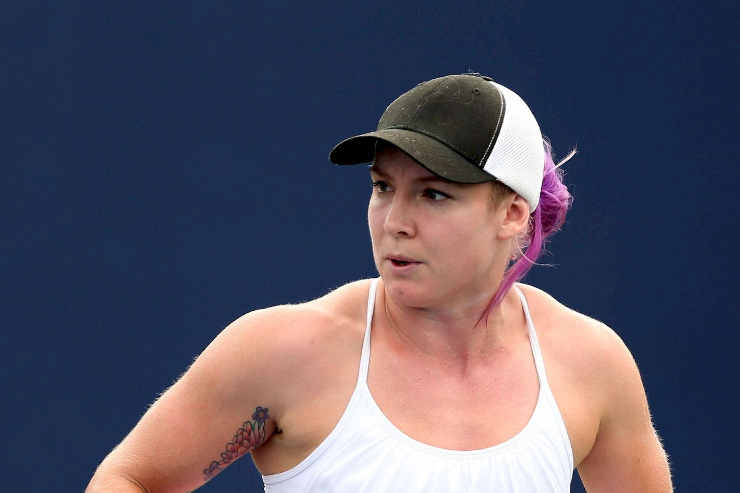 Tennis 桌布 壁紙 — 一位專注的女性網球運動員，紫色頭髮和刺青，戴著帽子，穿著白色背心，背景為純藍色。