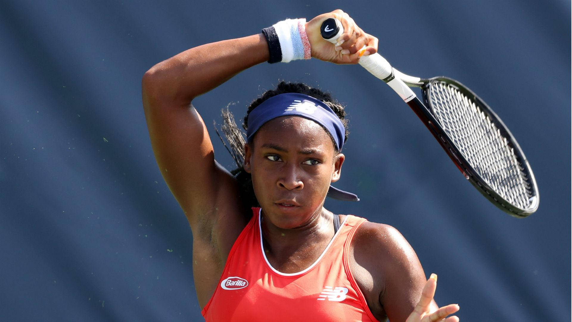 Coco Gauff 桌布 壁紙 — 一位專注的女性網球選手穿著紅色上衣，準備在純藍色背景前發球。