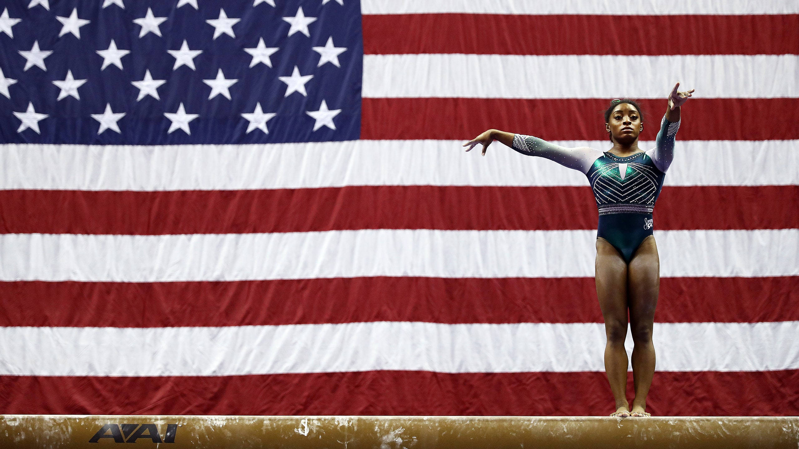 simon biles 桌布 壁紙 — 一位專注的女性體操運動員在平衡木上伸展雙臂，背景是大型美國國旗。