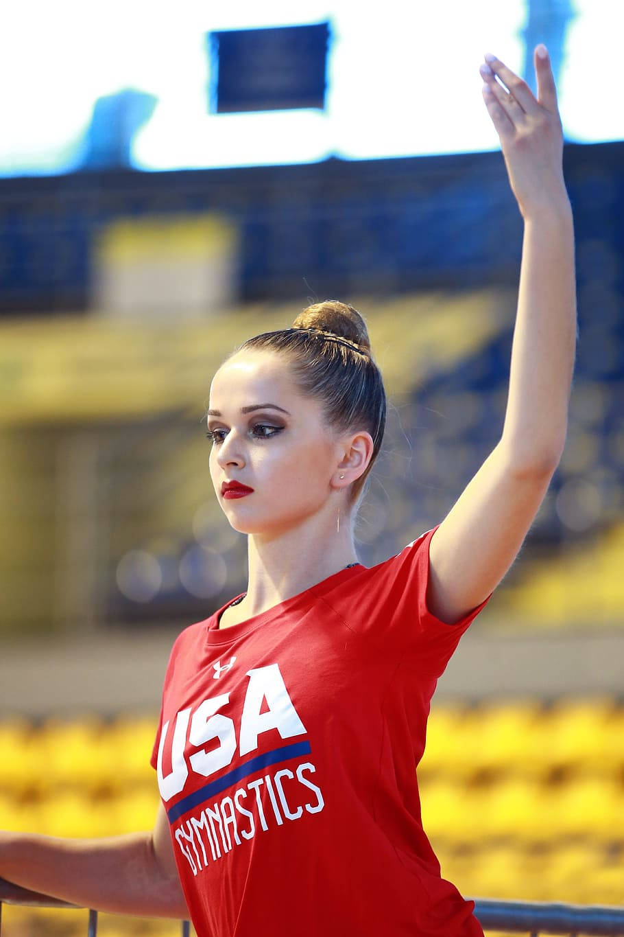 Athletic 桌布 壁紙 — 一位專注的女性體操運動員穿著紅色USA Gymnastics襯衫，在明亮的室內競技場中舉起手臂，場內有黃色座位。