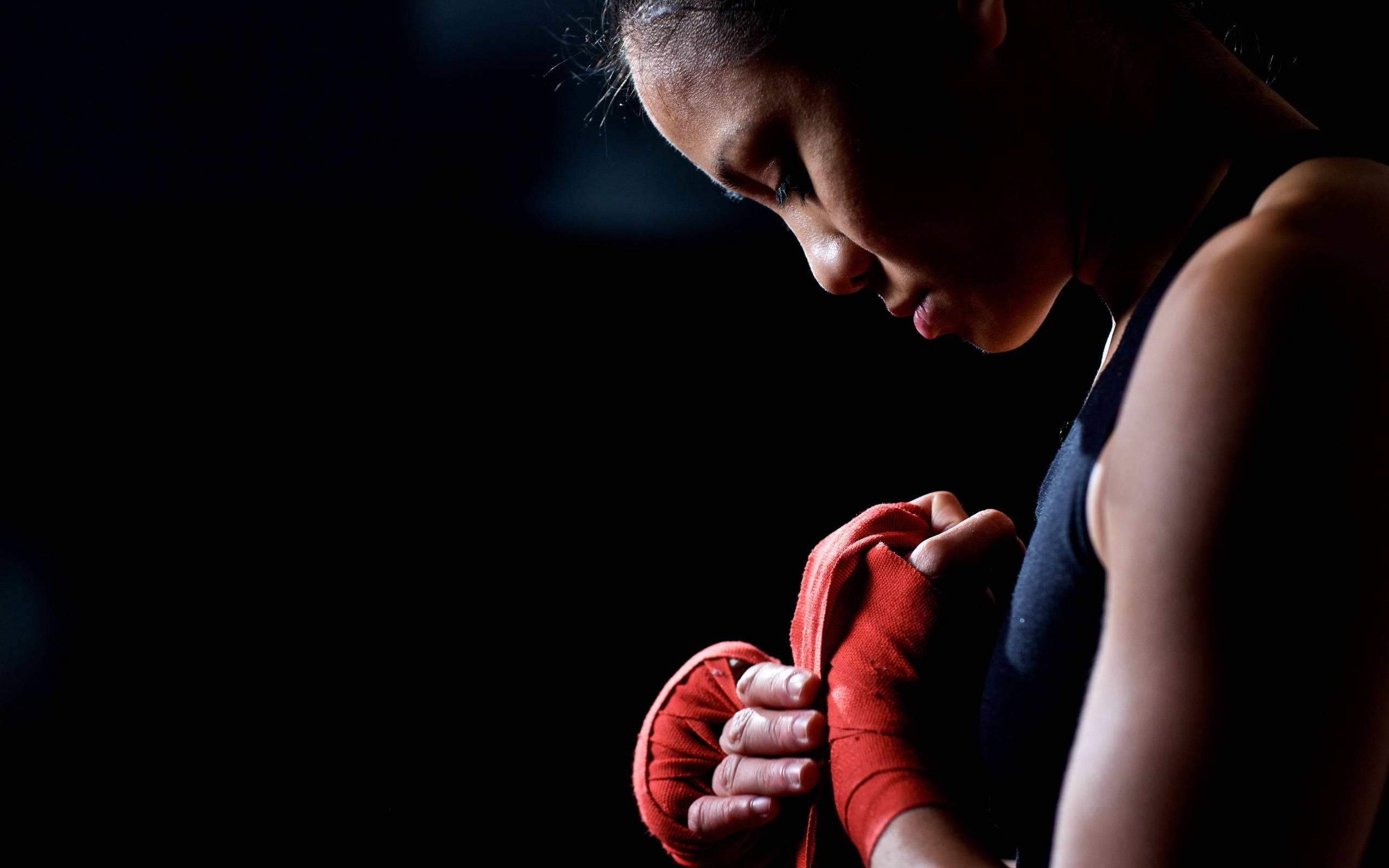 Boxing 桌布 壁紙 — 一位專注的女戰士在昏暗燈光下，帶著強烈的決心在比賽前纏繞紅色護手帶。