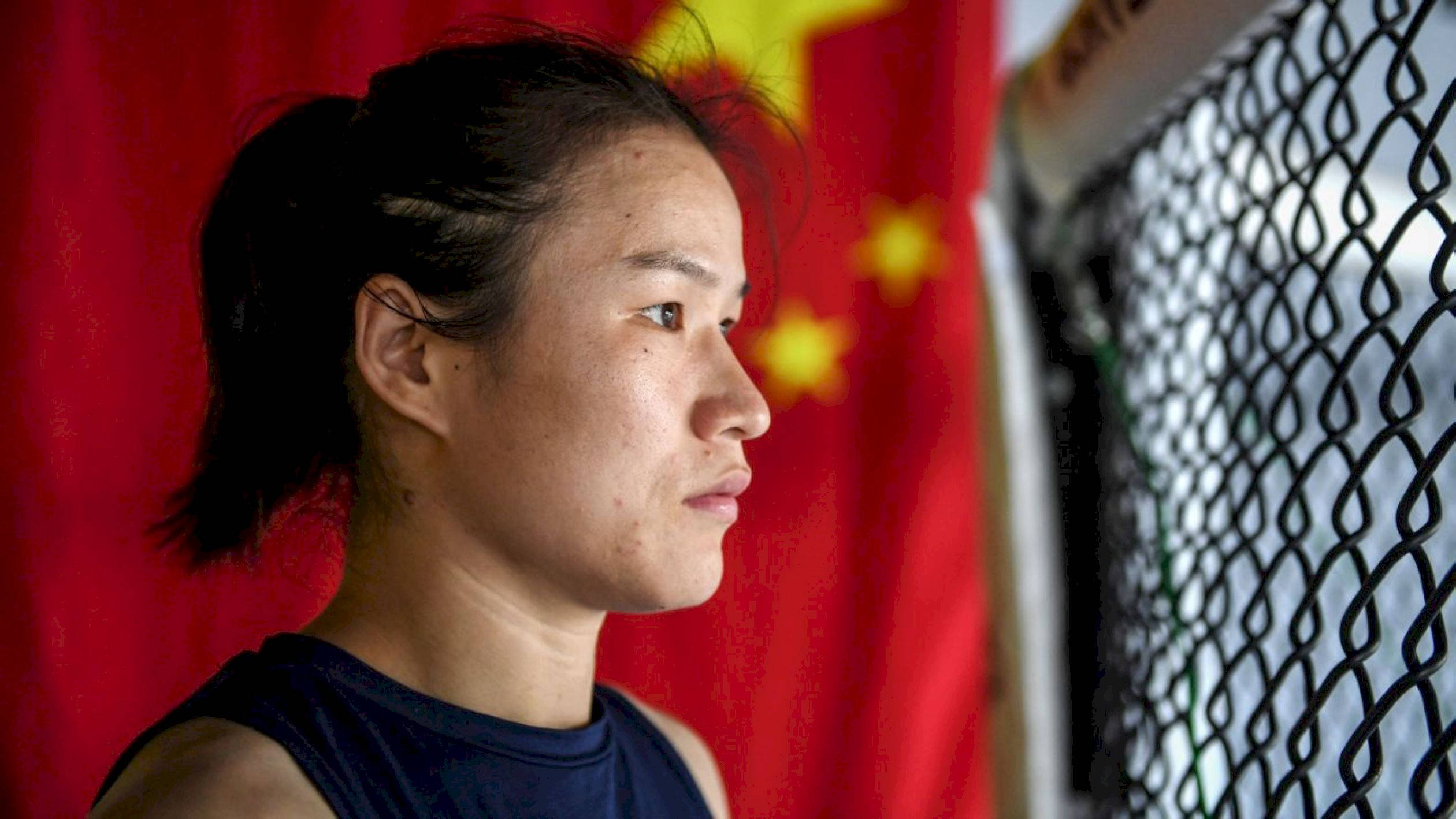 MMA 桌布 壁紙 — 一位專注的女戰士身穿藍色背心，透過鐵絲網凝視，身後有中國國旗。