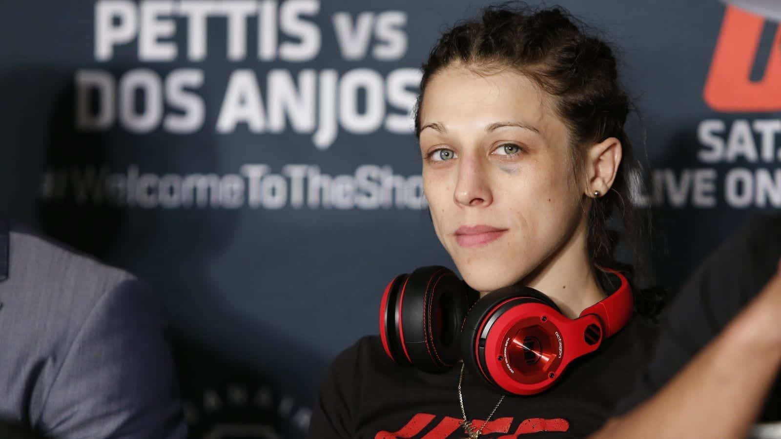 professional 桌布 壁紙 — 一位專注的女拳手，脖子上掛著紅色耳機，在 'Pettis vs Dos Anjos' 背景的 UFC 記者會上擺姿勢。
