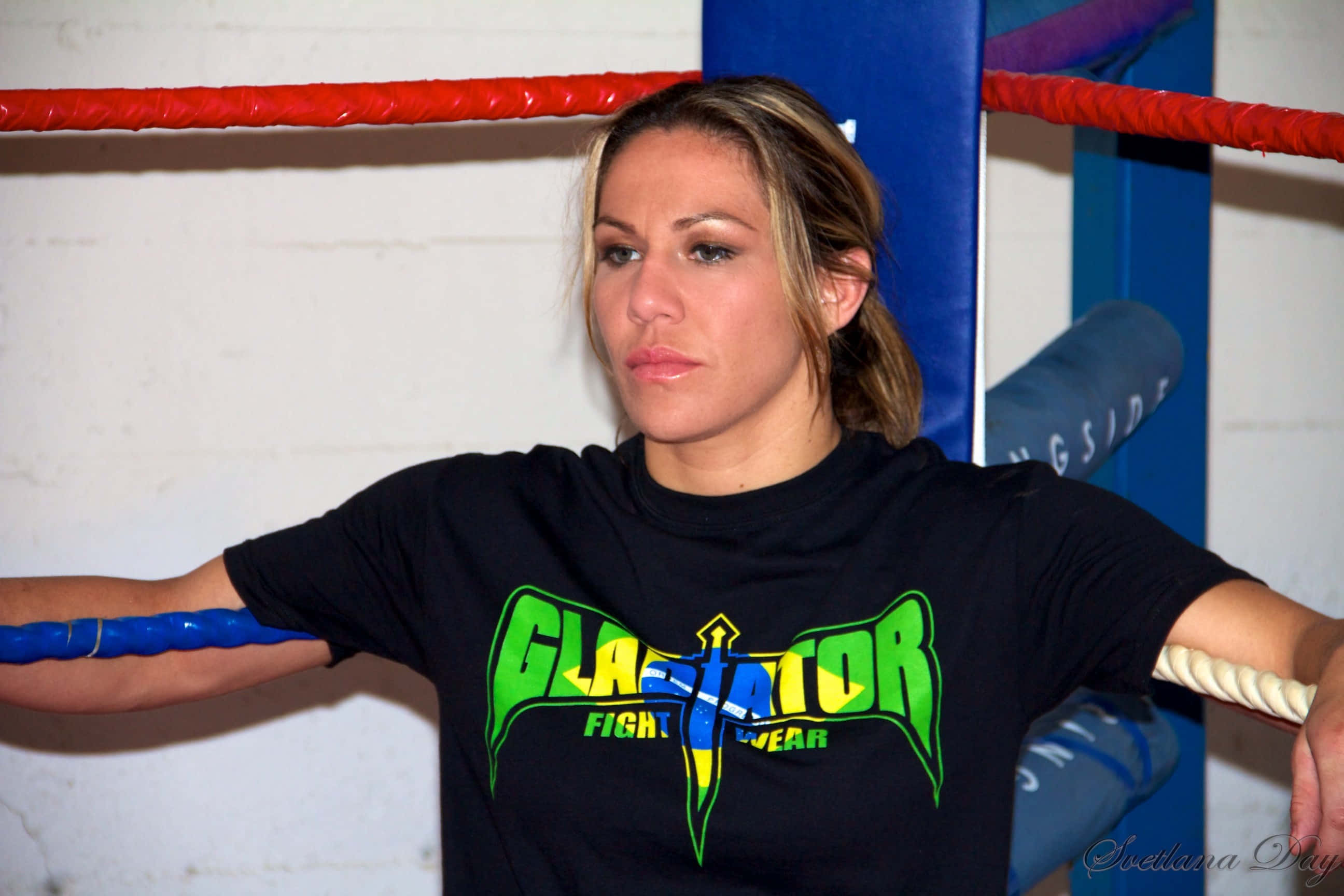 UFC 桌布 壁紙 — 一位專注的女拳手靠在拳擊繩上，身穿黑色「Gladiator Fight Wear」T恤。