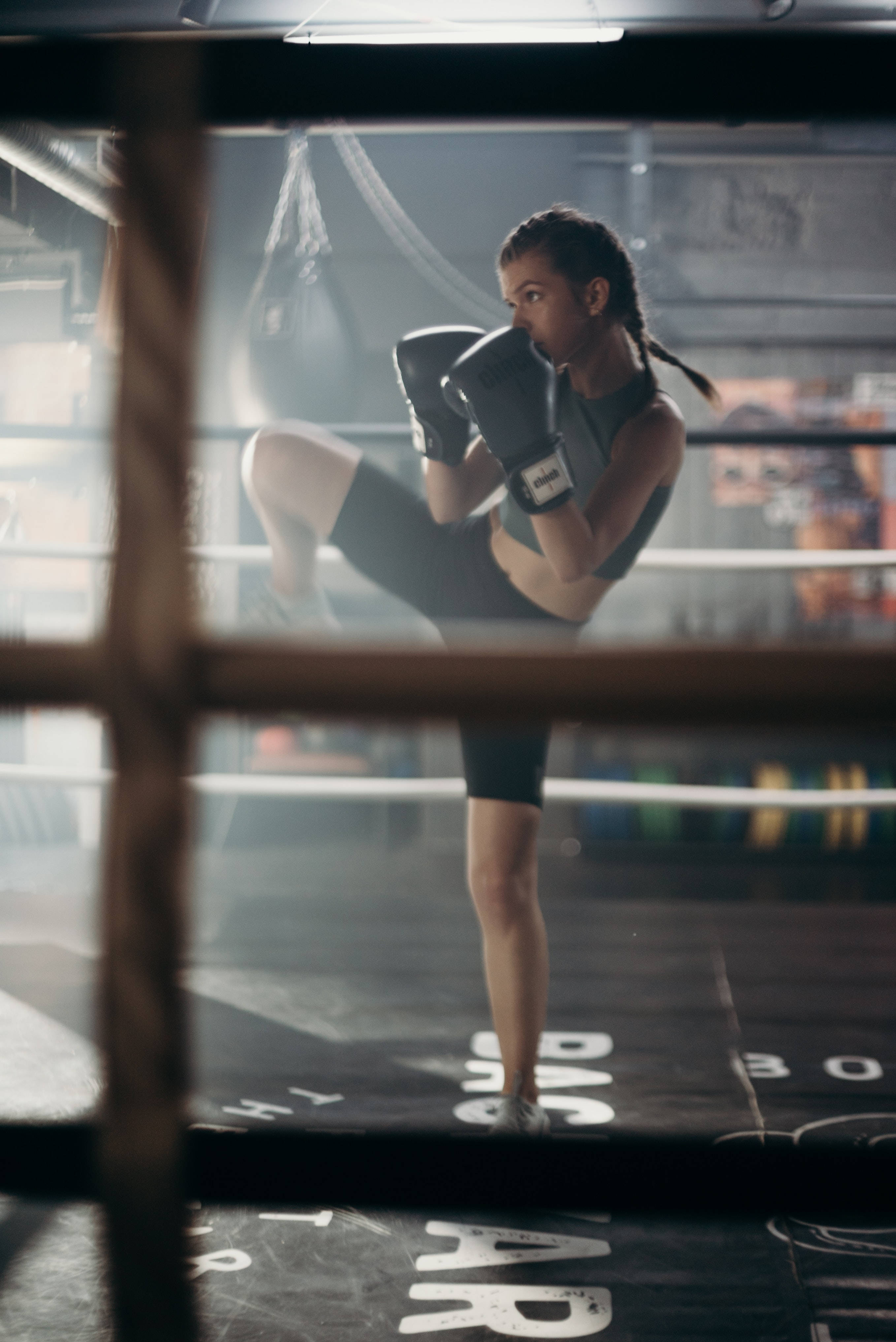 Cool Boxing 桌布 壁紙 — 一位專注的女拳擊手在煙霧瀰漫的拳擊擂台上，被繩索和昏暗的頂燈框住，施展高踢腿。