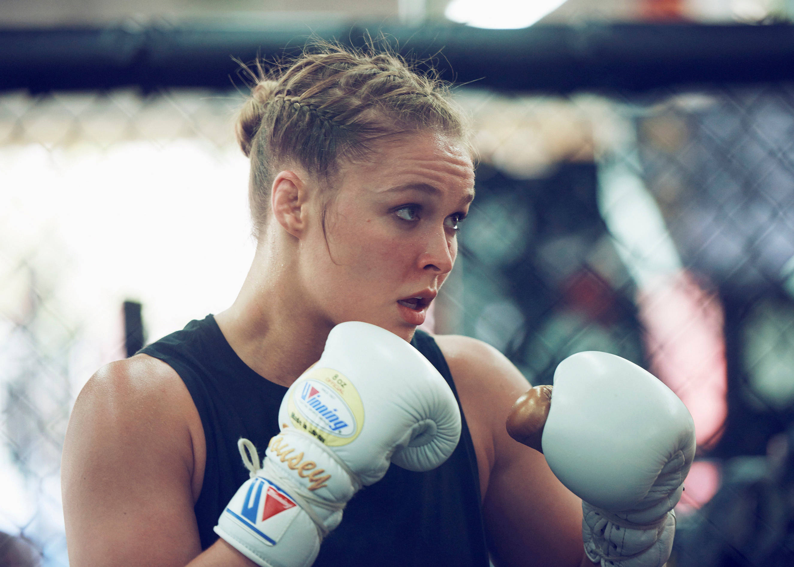 Rousey 桌布 壁紙 — 一位專注的女拳擊手戴著白色手套，在模糊的健身房背景下於籠內進行高強度訓練。