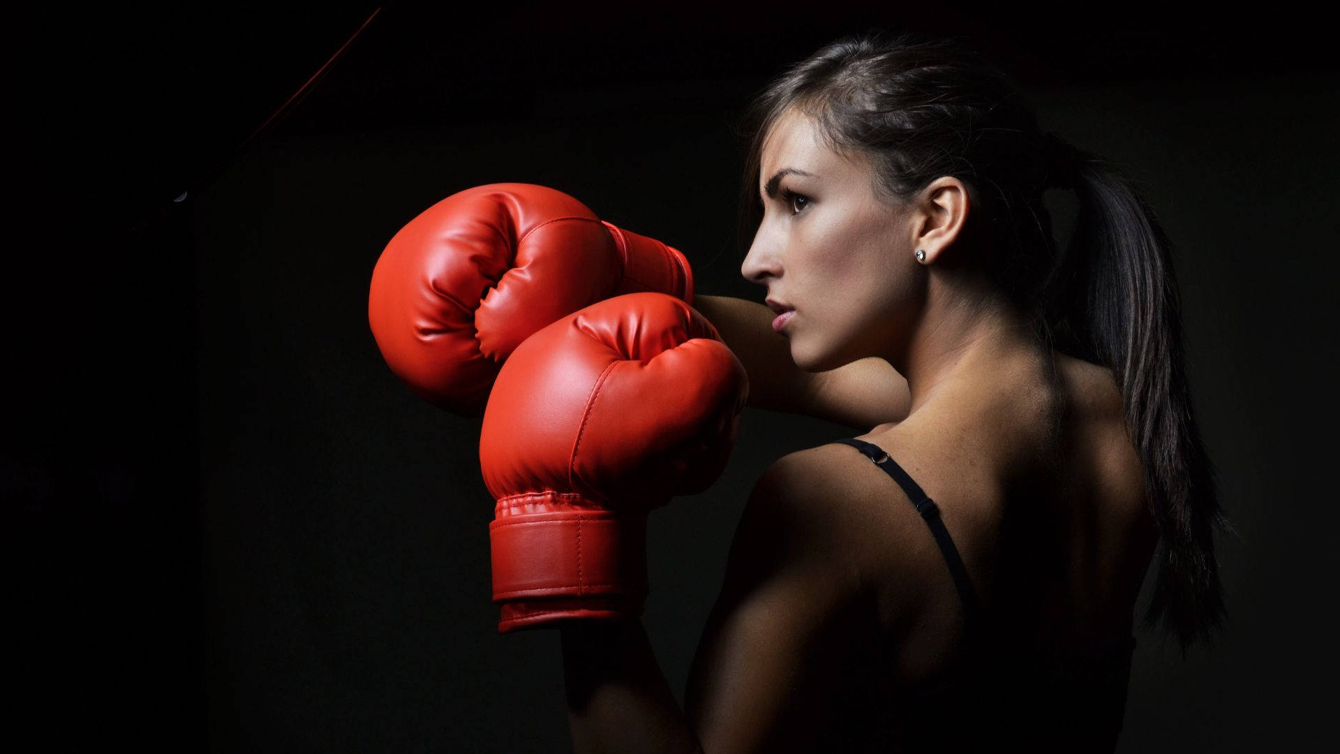 Boxing 桌布 壁紙 — 一位專注的女拳擊手戴著紅色手套，在黑暗背景下被戲劇性的燈光照亮，擺好姿勢。