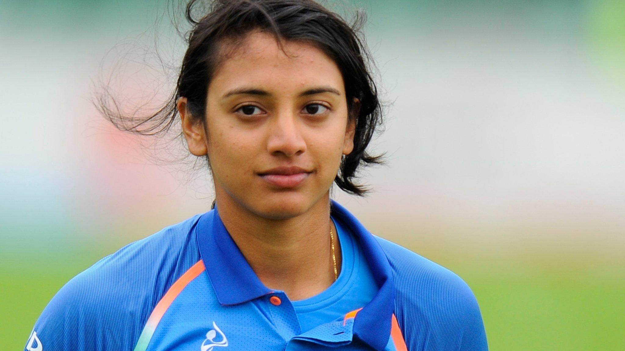 Smriti Mandhana 桌布 壁紙 — 一位專注的女板球運動員身穿藍色球衣，站在戶外，風吹亂了她的黑髮，背景是模糊的綠色球場。