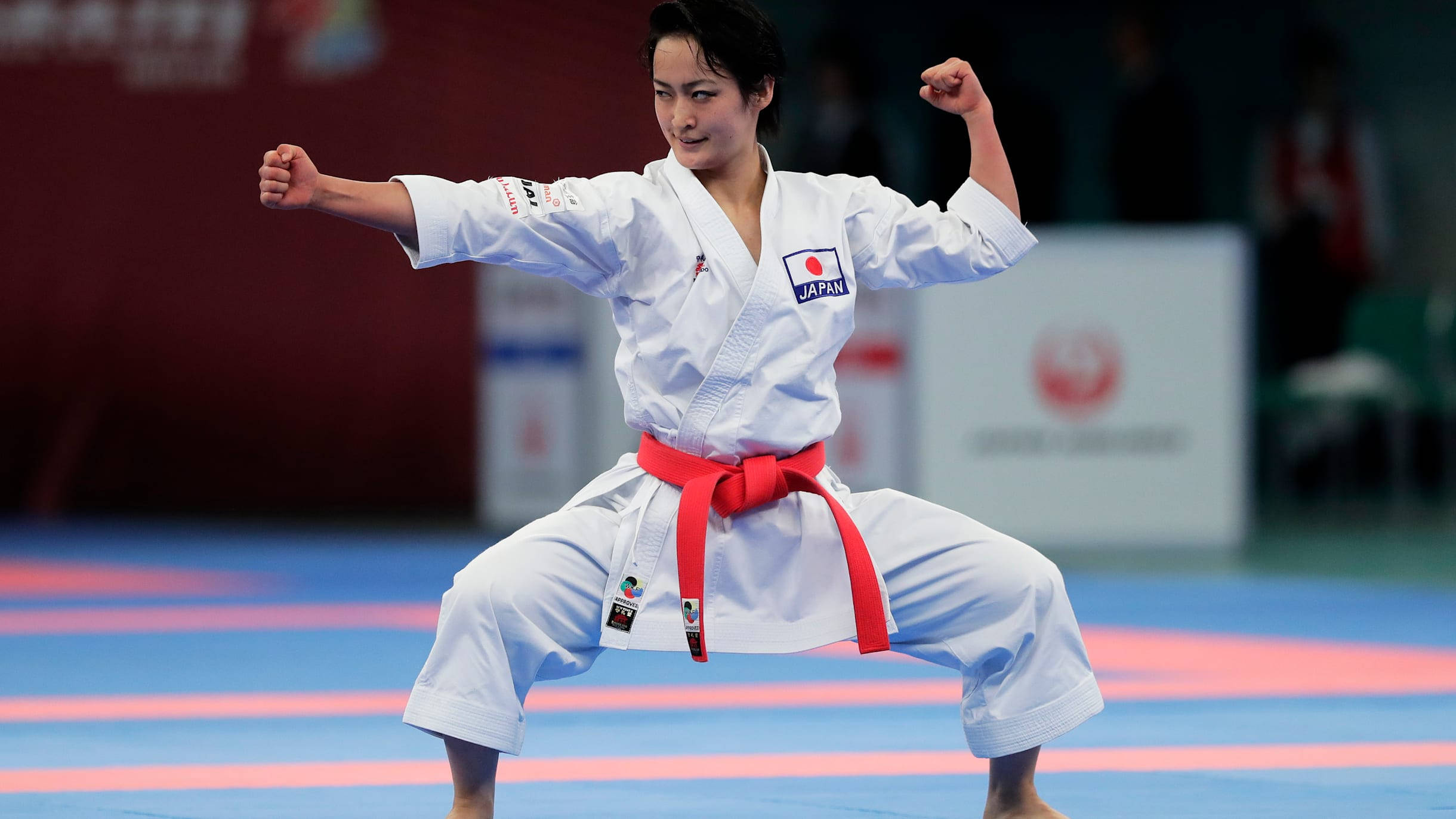 Athletic 桌布 壁紙 — 一位專注的女空手道選手，身穿白色道服、繫紅色腰帶，在藍色比賽墊上擺出強而有力的姿勢。