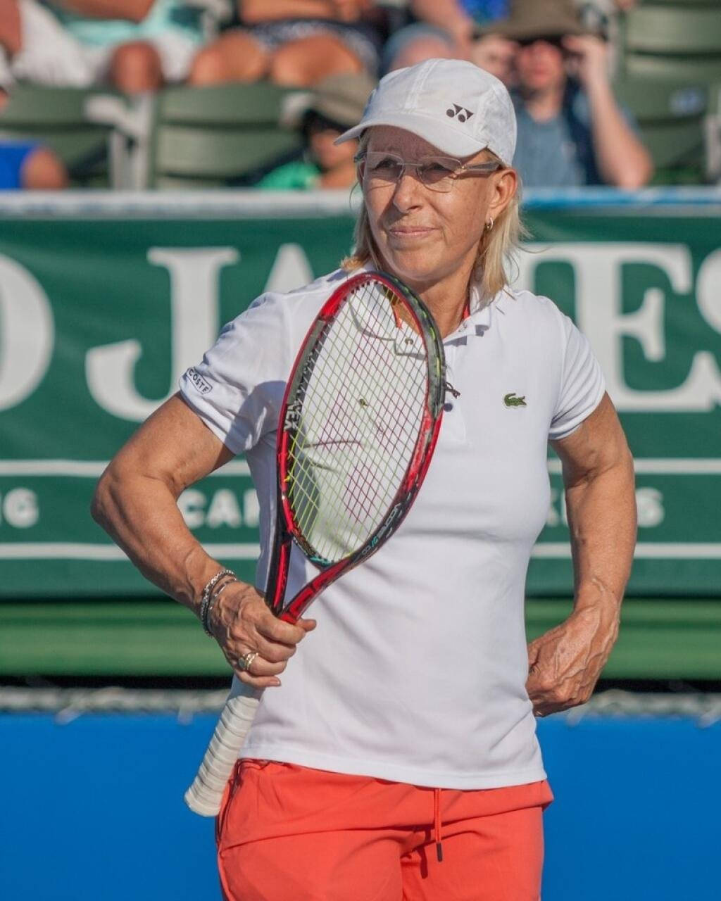 Martina Navratilova 桌布 壁紙 — 一位專注的女網球運動員，戴著白色帽子，穿著紅色短褲，在藍色球場上握著球拍，身後有觀眾坐在綠色座位上。