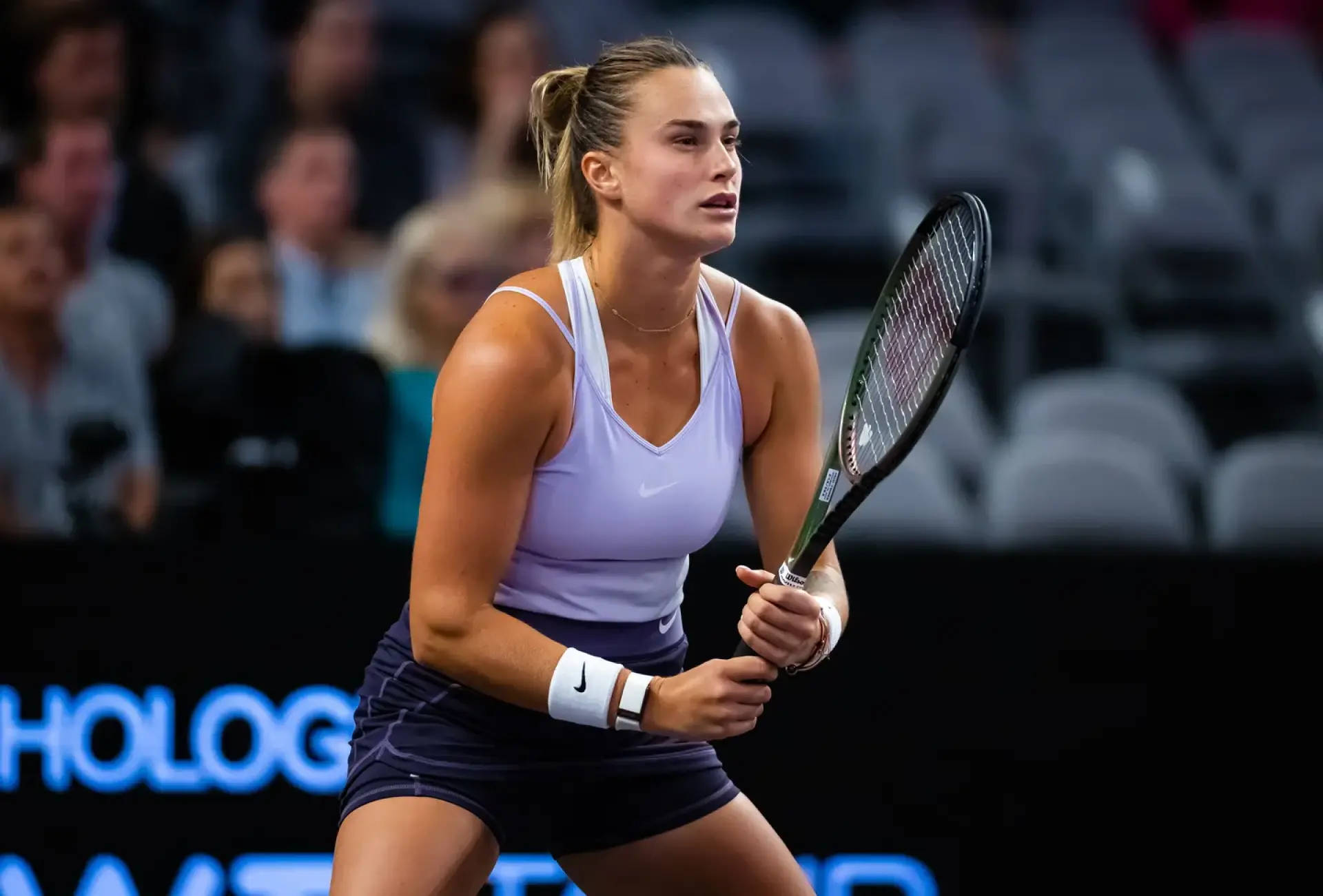 Aryna Sabalenka 桌布 壁紙 — 一位專注的女網球運動員，身穿薰衣草色上衣和海軍藍裙子，在光線明亮的室內球場中握緊球拍進行比賽。