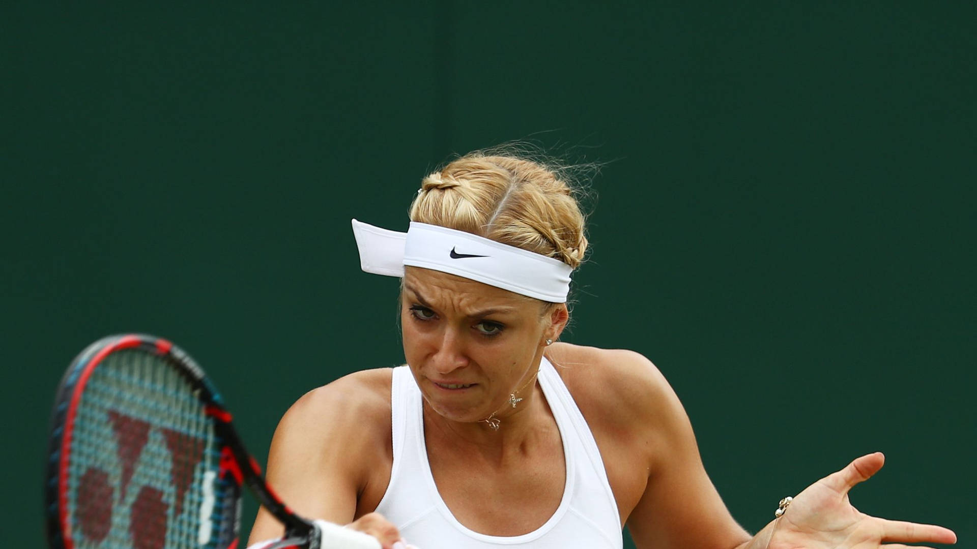 Tennis 桌布 壁紙 — 一位專注的女網球運動員，金髮，戴白色頭帶，在深綠色球場背景下揮拍中握緊球拍。