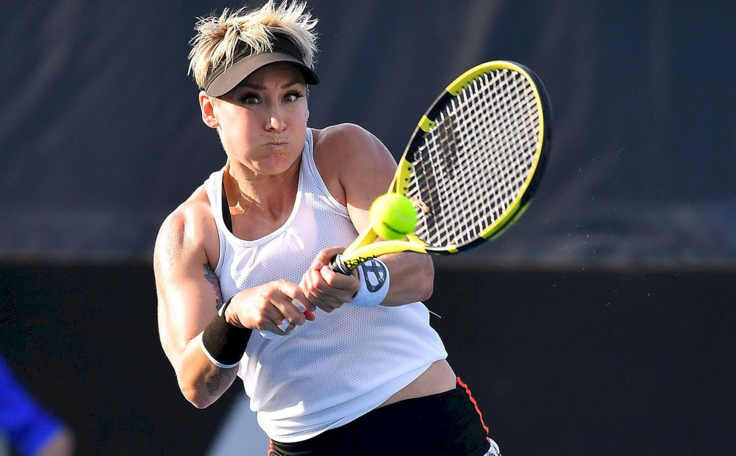 Tennis 桌布 壁紙 — 一位專注的女網球運動員在揮拍中擊打一顆亮黃色的球，使用黃黑相間的球拍，背景為深色。