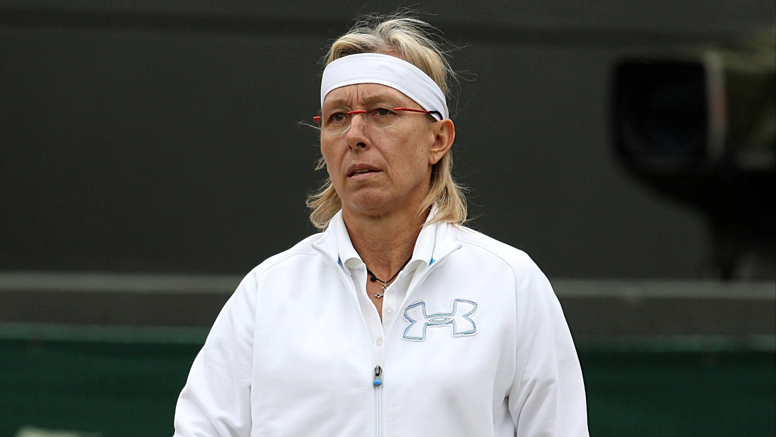 Martina Navratilova 桌布 壁紙 — 一位專注的女網球運動員穿著白色Under Armour外套和頭帶，帶著嚴肅的表情站在球場上。
