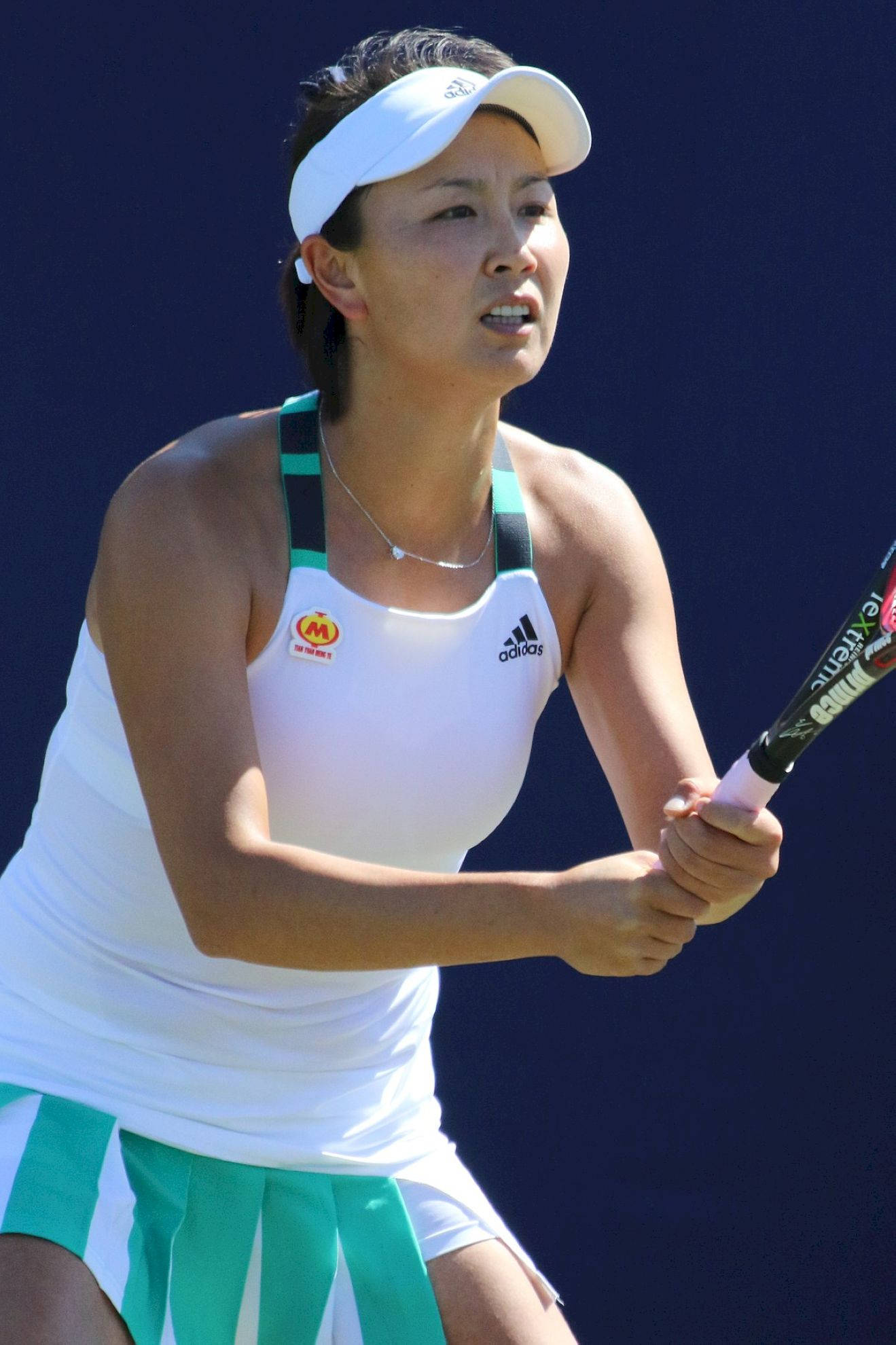 Tennis 桌布 壁紙 — 一位專注的女網球運動員穿著白色和綠色的裝備，在純藍色背景前揮拍中途緊握球拍。