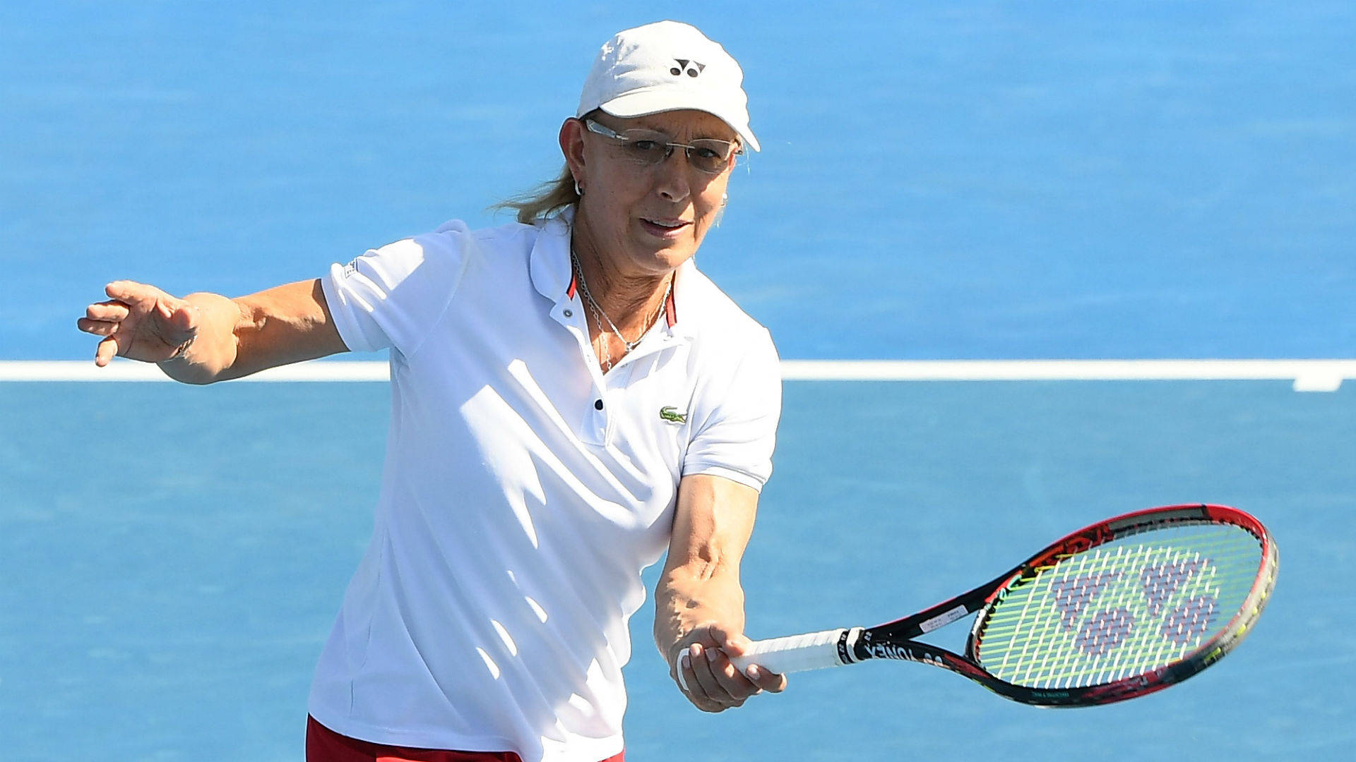 Martina Navratilova 桌布 壁紙 — 一位專注的女網球運動員穿著白色裝備，在明亮的藍色球場上，在清晰的日光下揮動球拍。