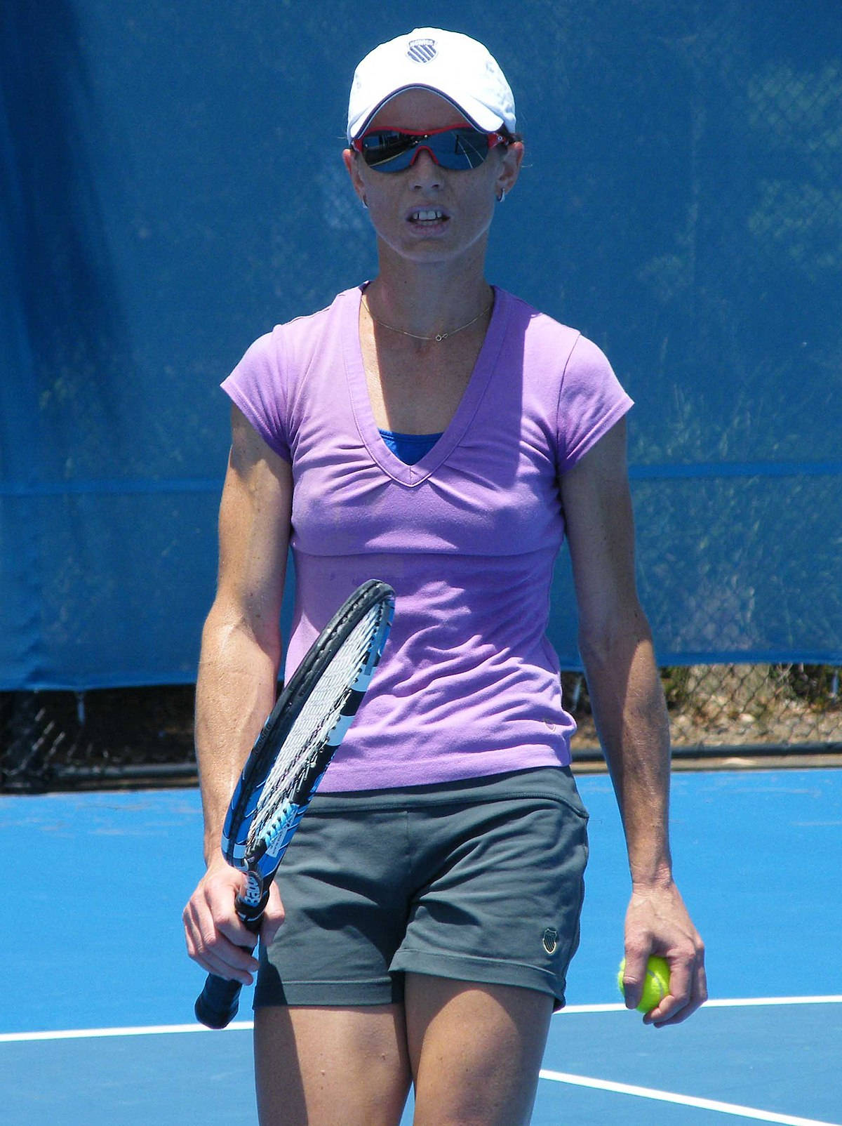 Tennis 桌布 壁紙 — 一位專注的女網球運動員穿著紫色襯衫和白色帽子，站在明亮的藍色球場上，手持球拍和球。