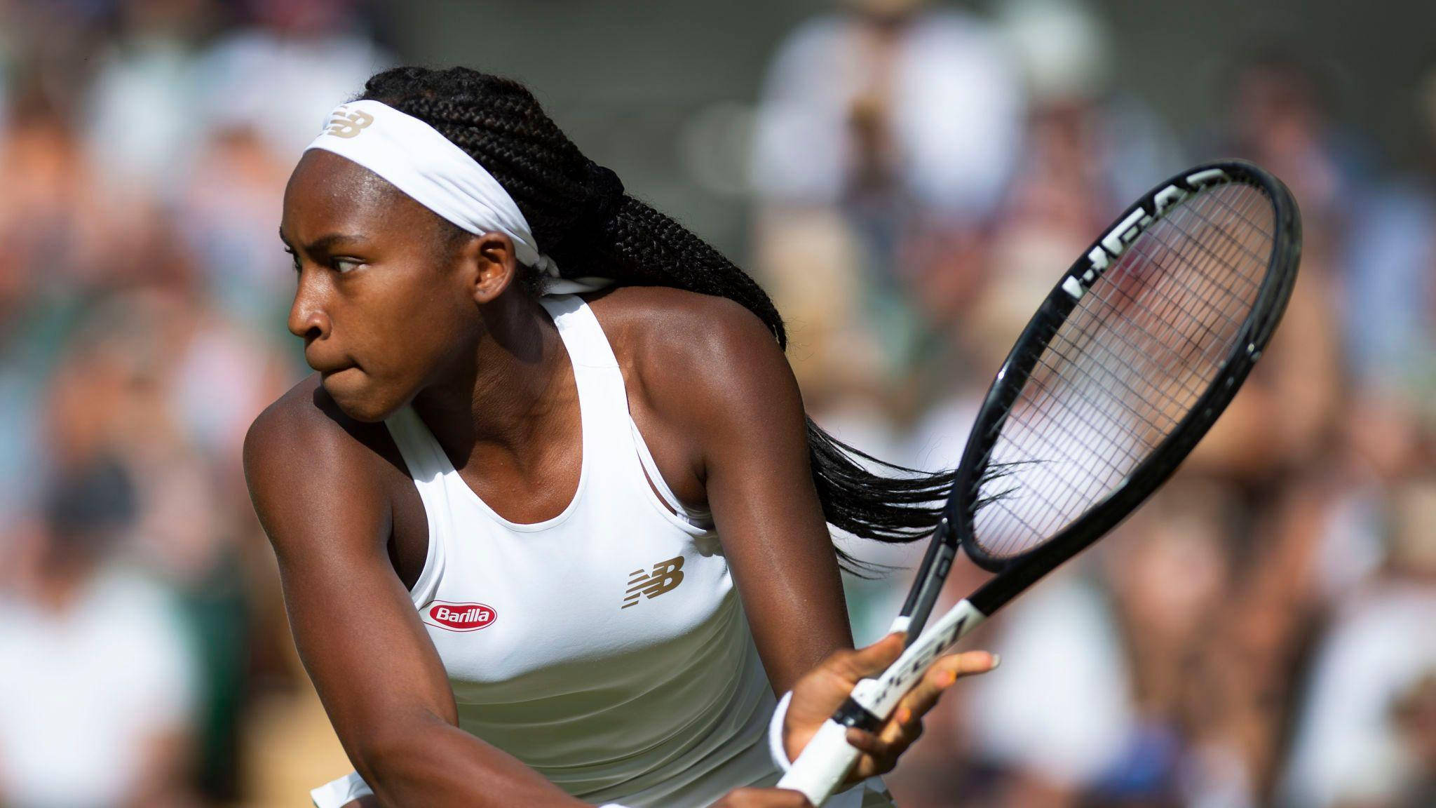 Coco Gauff 桌布 壁紙 — 一位專注的女網球運動員身穿白色裝備，在陽光照耀的草地場上揮拍中途握緊球拍，身後是模糊的觀眾。