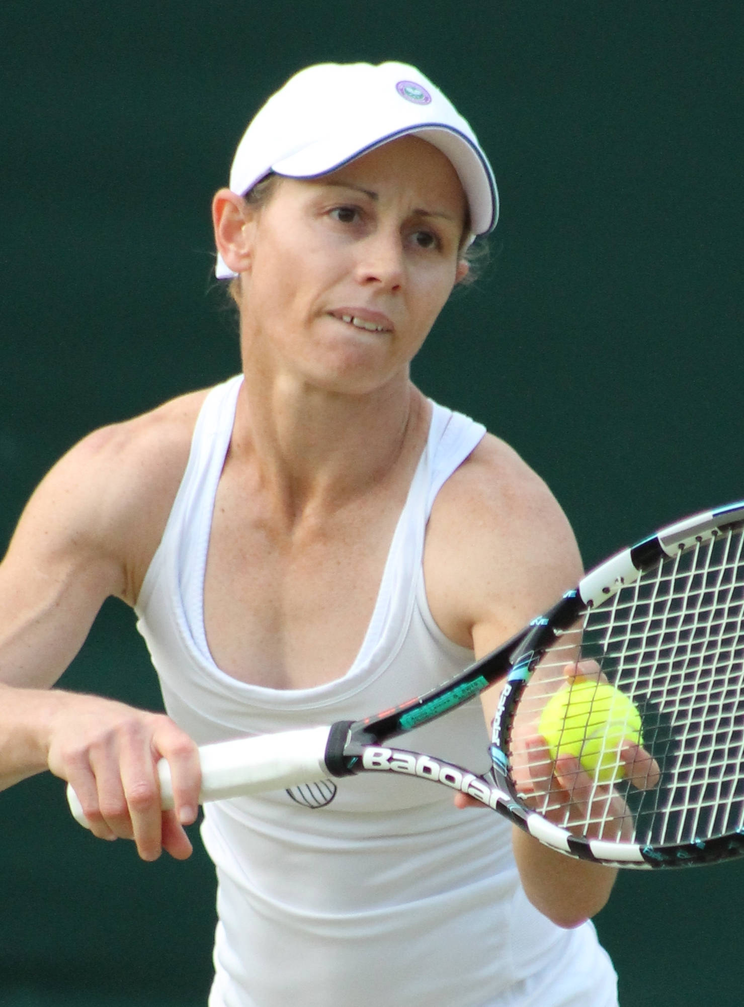 Tennis 桌布 壁紙 — 一位專注的女網球運動員身穿白色裝備，在深綠色球場背景下握著球拍和黃色球。