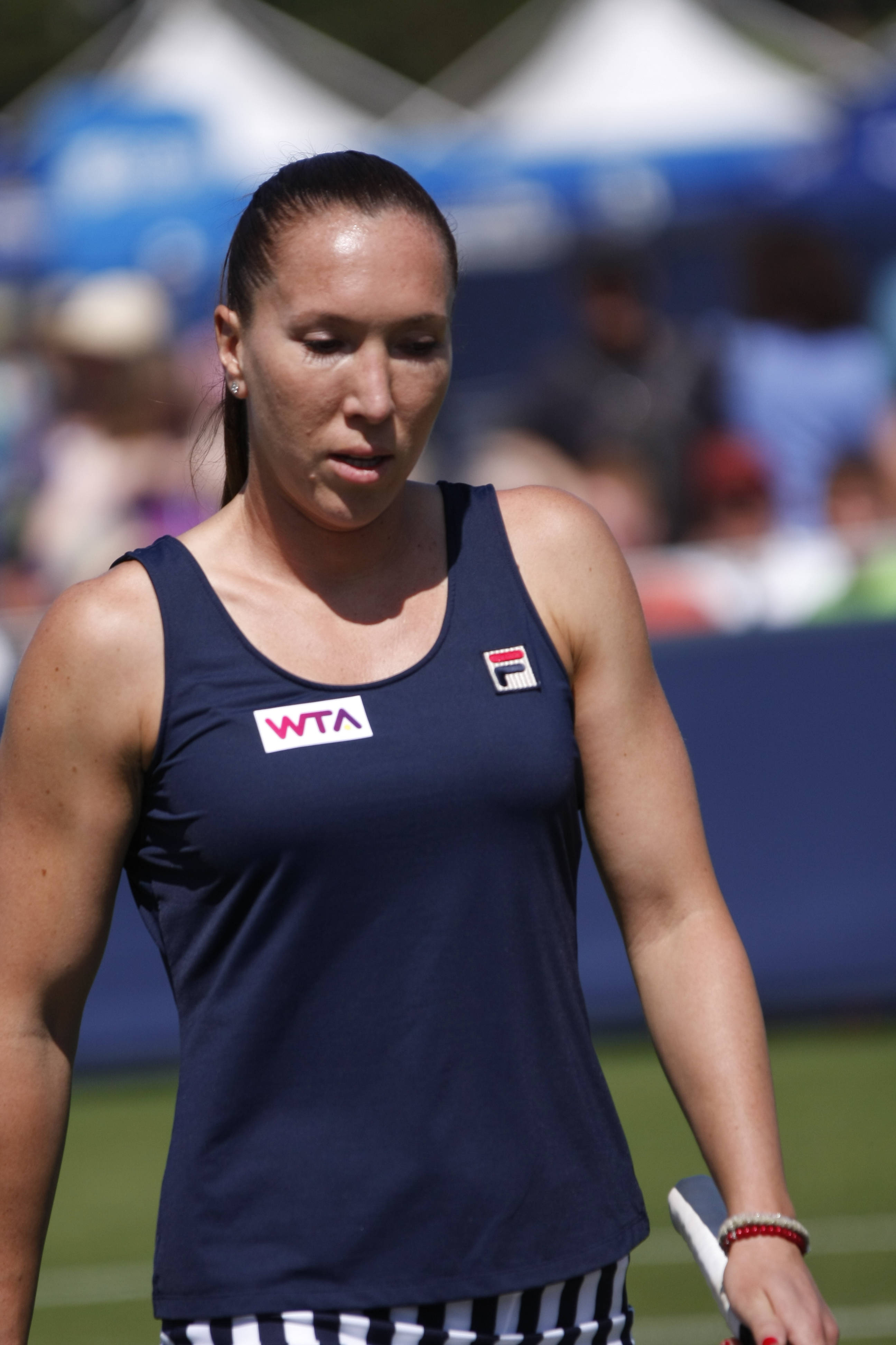 Tennis 桌布 壁紙 — 一位專注的女網球選手身穿海軍藍WTA背心，在陽光明媚的球場上行走，背景觀眾模糊。