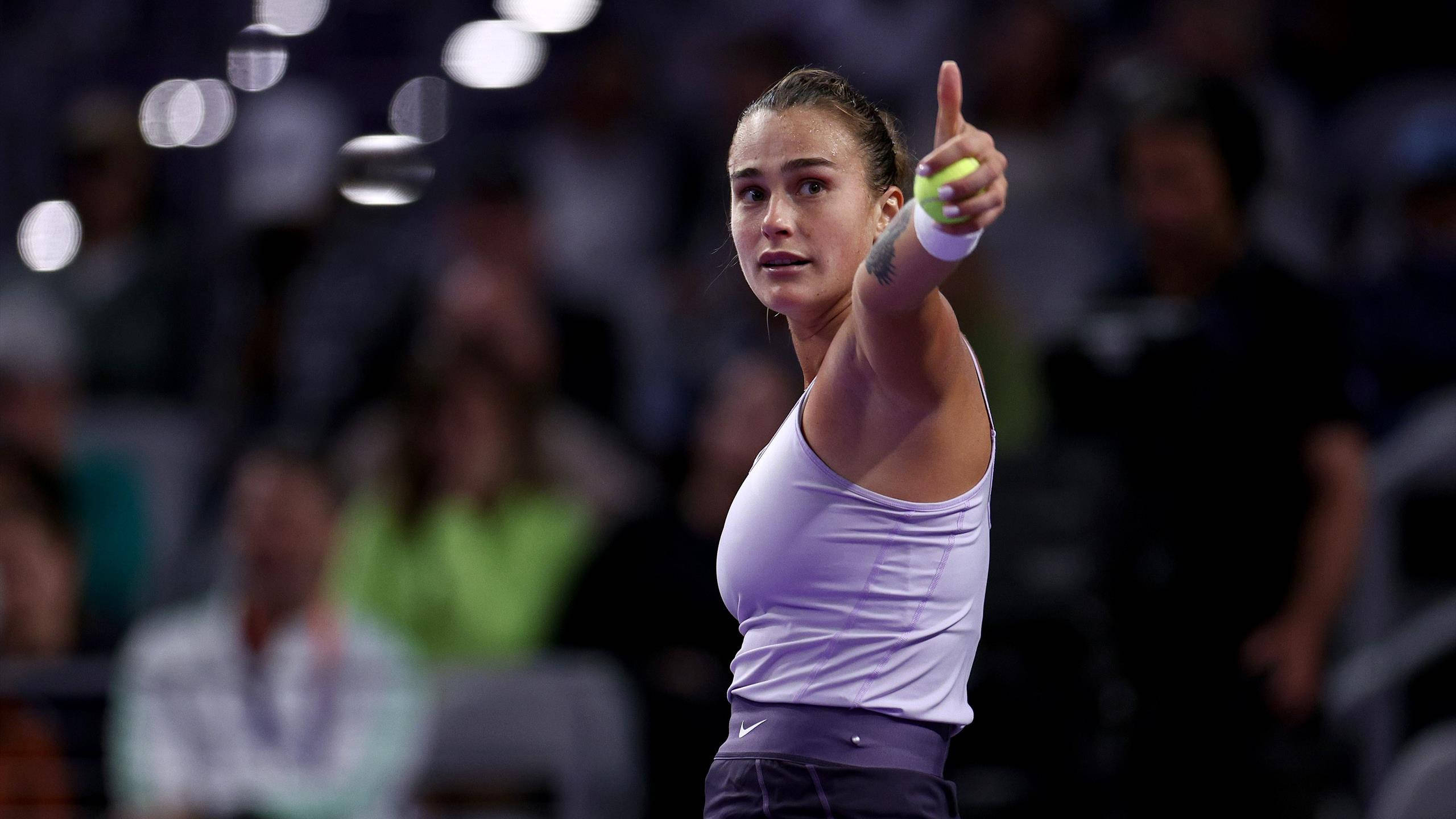 Aryna Sabalenka 桌布 壁紙 — 一位專注的女網球選手身穿白色上衣，在明亮的體育場燈光下，手持黃球準備發球。